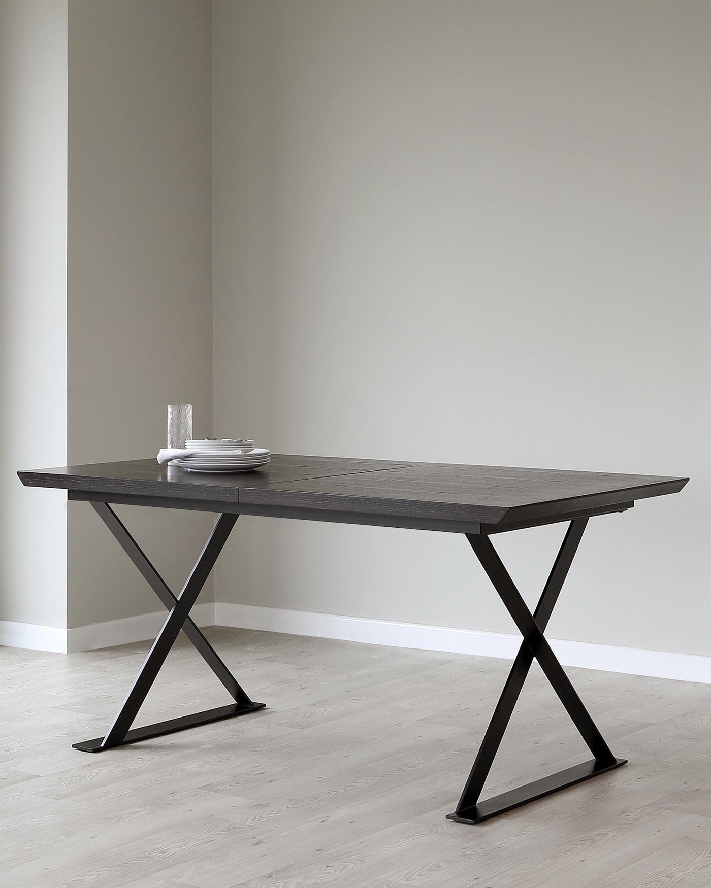 Modern minimalist dining table featuring a dark wood finish tabletop and sleek, angular black metal legs forming an X shape. The table is styled with a simple set of white plates, showcasing its clean lines and contemporary design.