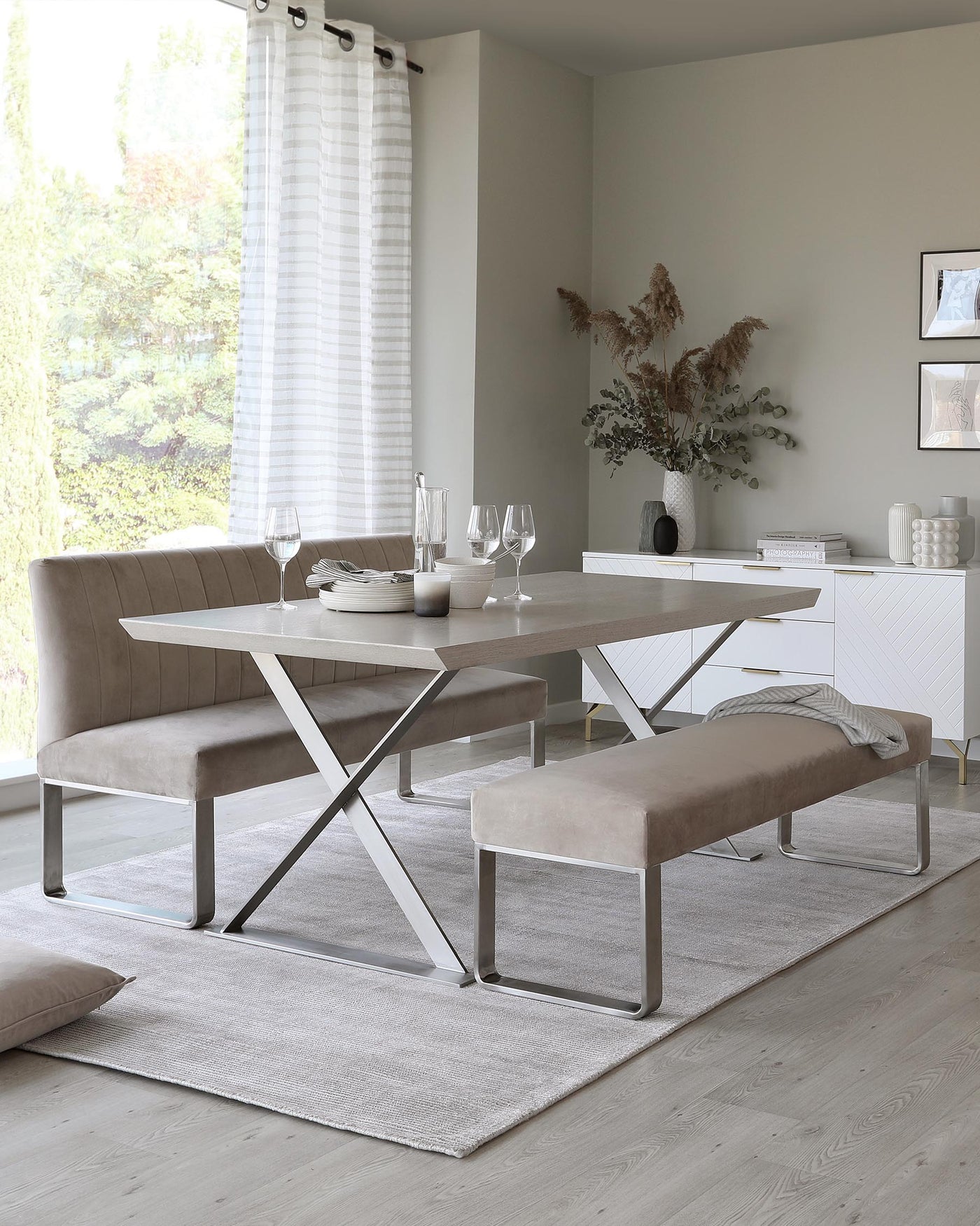 Modern dining room furniture set including a rectangular table with a white marble top and distinctive crisscross silver metal legs. Accompanying the table is a taupe upholstered L-shaped bench and a matching bench, both with sleek silver metal frames. The set is arranged on a light grey area rug on a wooden floor, with a minimalist sideboard and decor in the background.