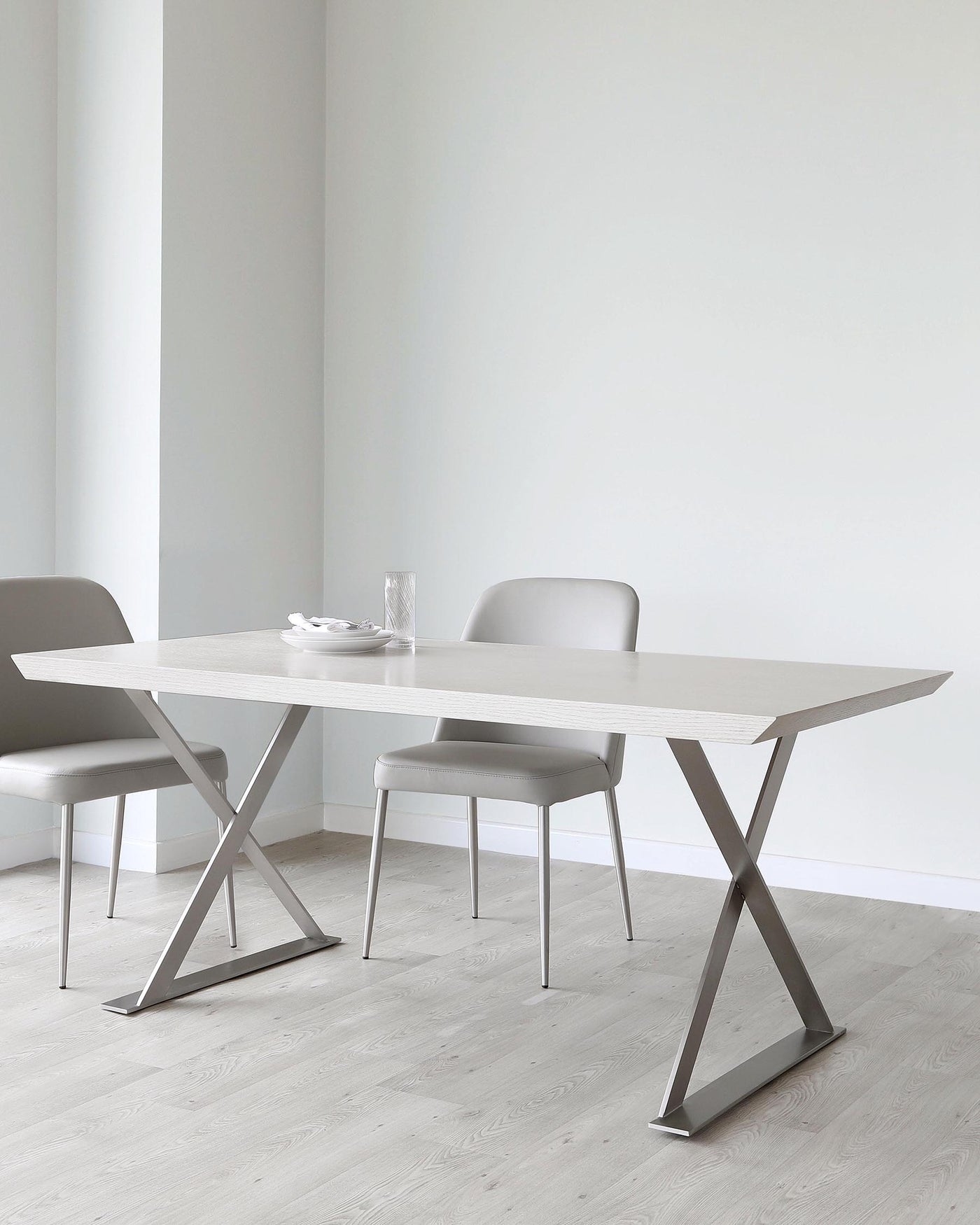 A modern minimalist dining set featuring a rectangular table with a light wood grain finish and distinctive X-shaped metallic legs. Two sleek chairs with padded seats and backrests, upholstered in a taupe fabric, complement the table, standing on slender metal legs. The set is displayed in a room with light grey walls and pale wooden flooring, emphasizing a clean and contemporary aesthetic.