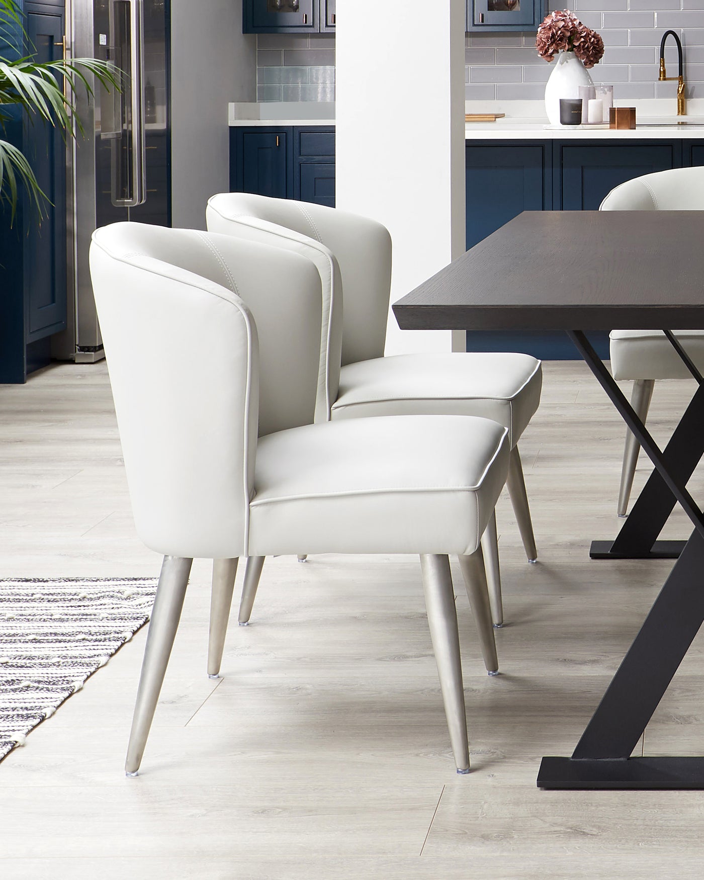 Elegant modern dining set featuring a dark wood table with black metal legs and a set of three sleek, light cream upholstered chairs with gently curved backs and angled metal legs. The scene is set against a contemporary kitchen backdrop with a blue cabinet finish and is complemented by a patterned rug on a light wooden floor.