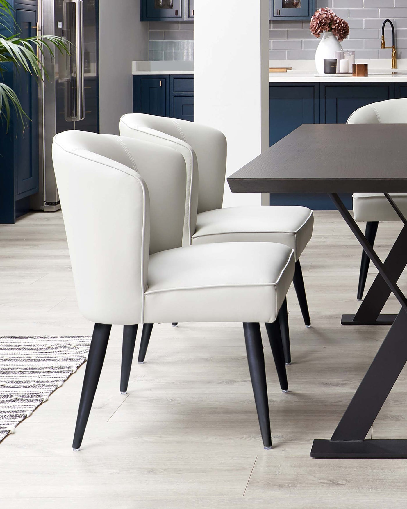 Modern dining room with a dark wooden rectangular table and a set of three sleek, off-white upholstered chairs with dark legs. The chairs have a curved backrest and plush seating. The setting is complemented by a contemporary kitchen in the background.