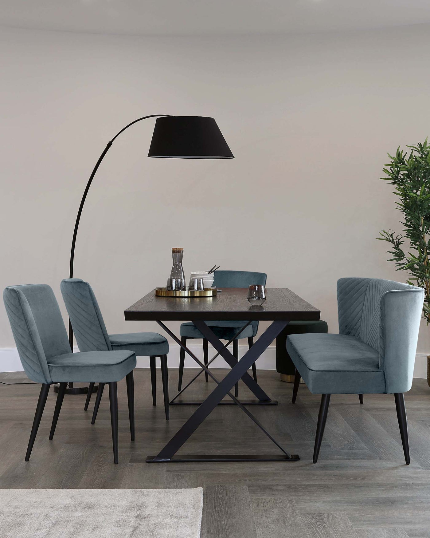 Modern dining set featuring a rectangular dark wood table with an X-shaped metal base and four plush velvet upholstered chairs in a soft blue colour, with channel tufting and black metal legs, complemented by an overhanging floor lamp with a large black shade.