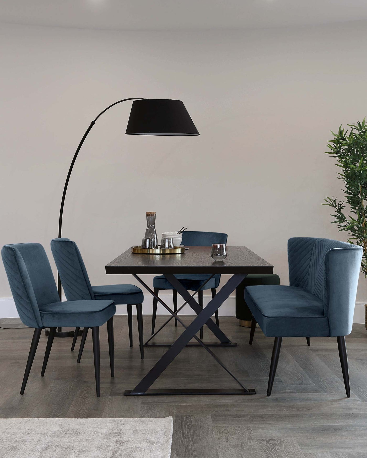 A modern dining room setting featuring a dark wooden rectangular table with an X-shaped metallic base. Around the table are four plush, blue velvet dining chairs with backrest quilting and slim, black metal legs. An overhanging floor lamp with a large, black shade creates a focused lighting effect over the table. A small green plant in a pot adds a touch of nature to the scene.