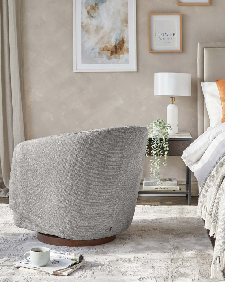 Contemporary grey fabric swivel chair with a rounded backrest, visible black zipper detailing, and a dark wood circular base, accompanied by a modern white table lamp on a glass-top side table with metallic accents. A draped throw blanket and decorative pillows are partially visible, suggesting a comfortable and stylish living space.