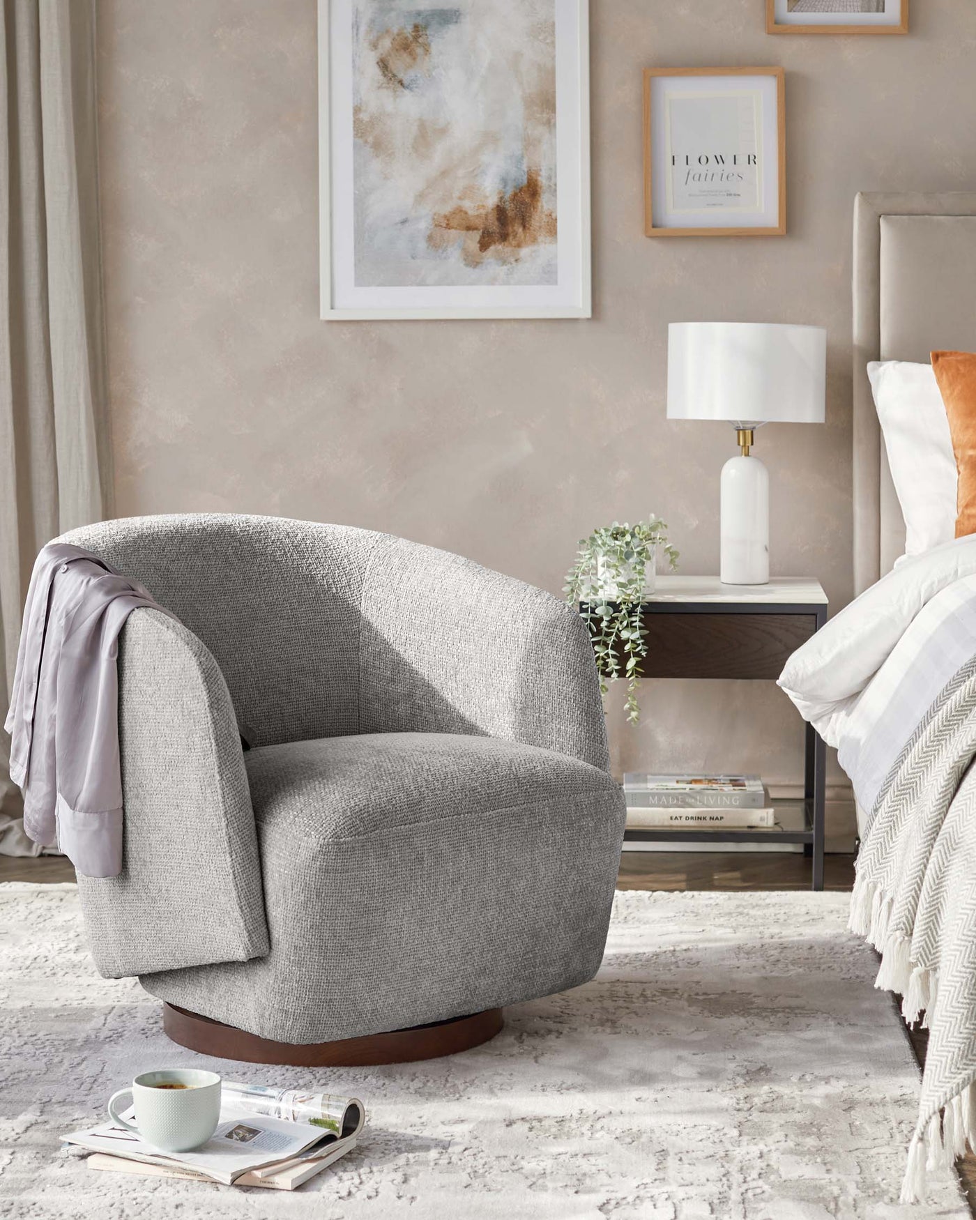 Contemporary plush grey fabric armchair with a curved back, wide seat, and dark wood base. Beside it, a sleek white side table with a lower shelf holding decorative items, topped with a modern white lamp with a cylindrical shade. An off-white textured throw is draped over the arm of the chair, and a light carpet beneath adds warmth to the scene.