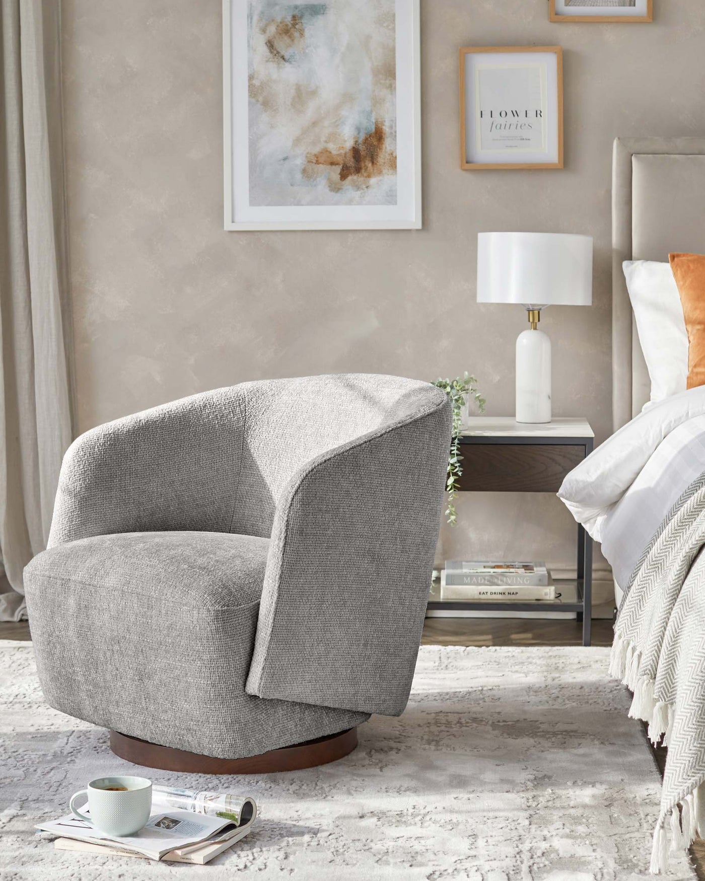 Modern textured grey fabric swivel chair with curved back and a deep, cushioned seat on a round, dark wooden base, paired with a sleek white cylindrical bedside lamp with a white shade on a minimalist bedside table with a reflective surface and lower shelf holding decorative items.
