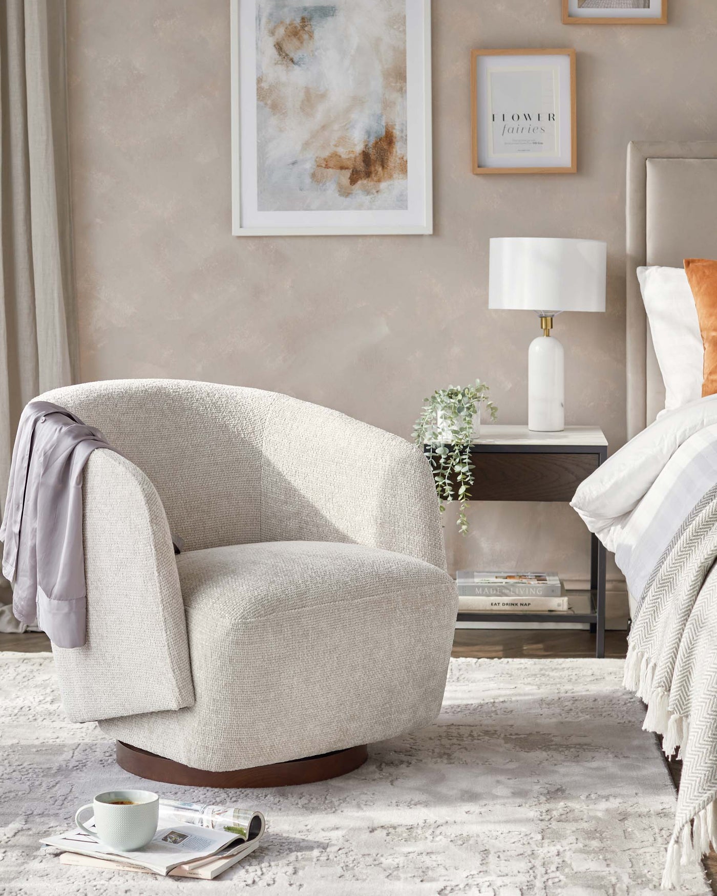 A contemporary beige upholstered swivel chair with a rounded design and a dark wooden base, accompanied by a modern white side table featuring a lower shelf, topped with a textured white lamp with a cylindrical shade. The setting includes a cosy beige throw blanket and a decorative plant, bringing warmth to the stylish interior.