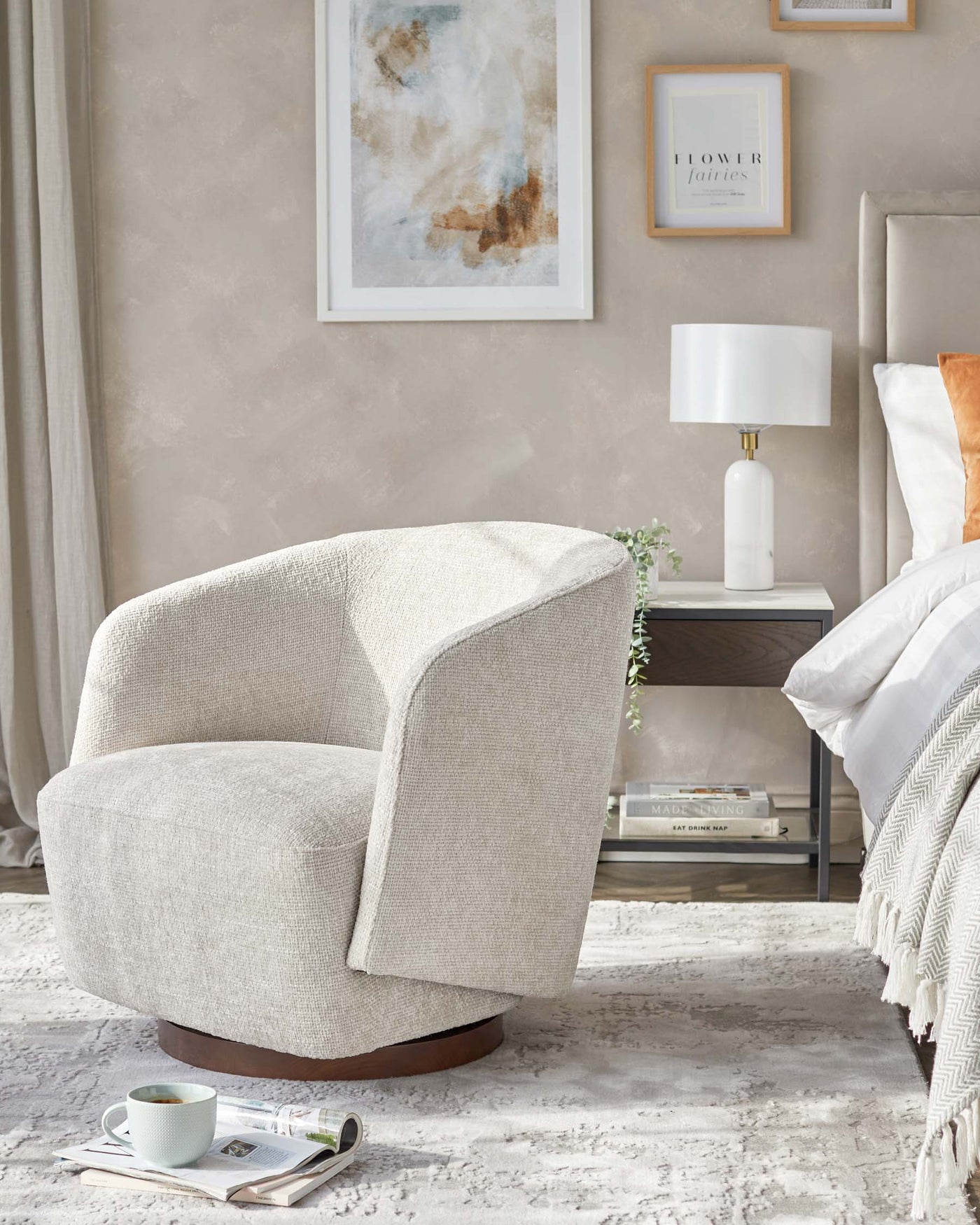 Modern stylish armchair with textured light beige upholstery and a unique curved design set on a dark wood base, beside a sleek white side table featuring a fashionable marble effect and metallic details, both adorned with minimalistic decor items and complementing the contemporary and cosy bedroom setup.