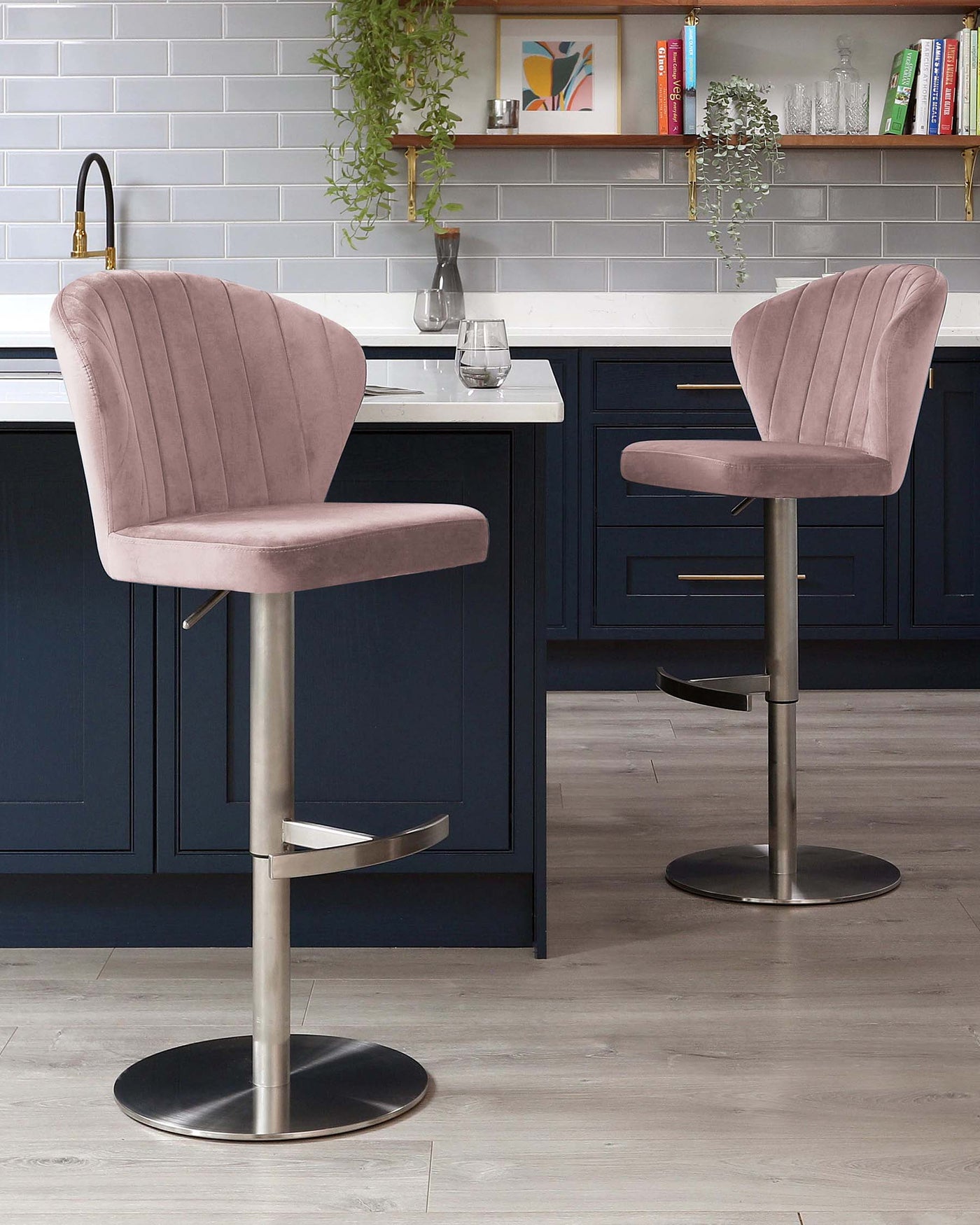 Two contemporary blush pink velvet bar stools with vertical channel tufting, curved backrests, and footrests on adjustable height brushed metal pedestal bases.