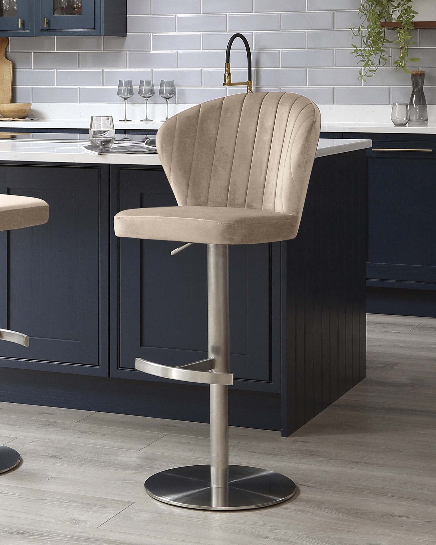 Elegant modern bar stool with velvet upholstery in a soft taupe colour, featuring vertical stitching on the curved backrest and a comfortable padded seat. The stool has a sleek metallic base with a circular footrest, adjustable height lever, and a sturdy round base, set against a kitchen island background with blue subway tile and dark cabinetry.