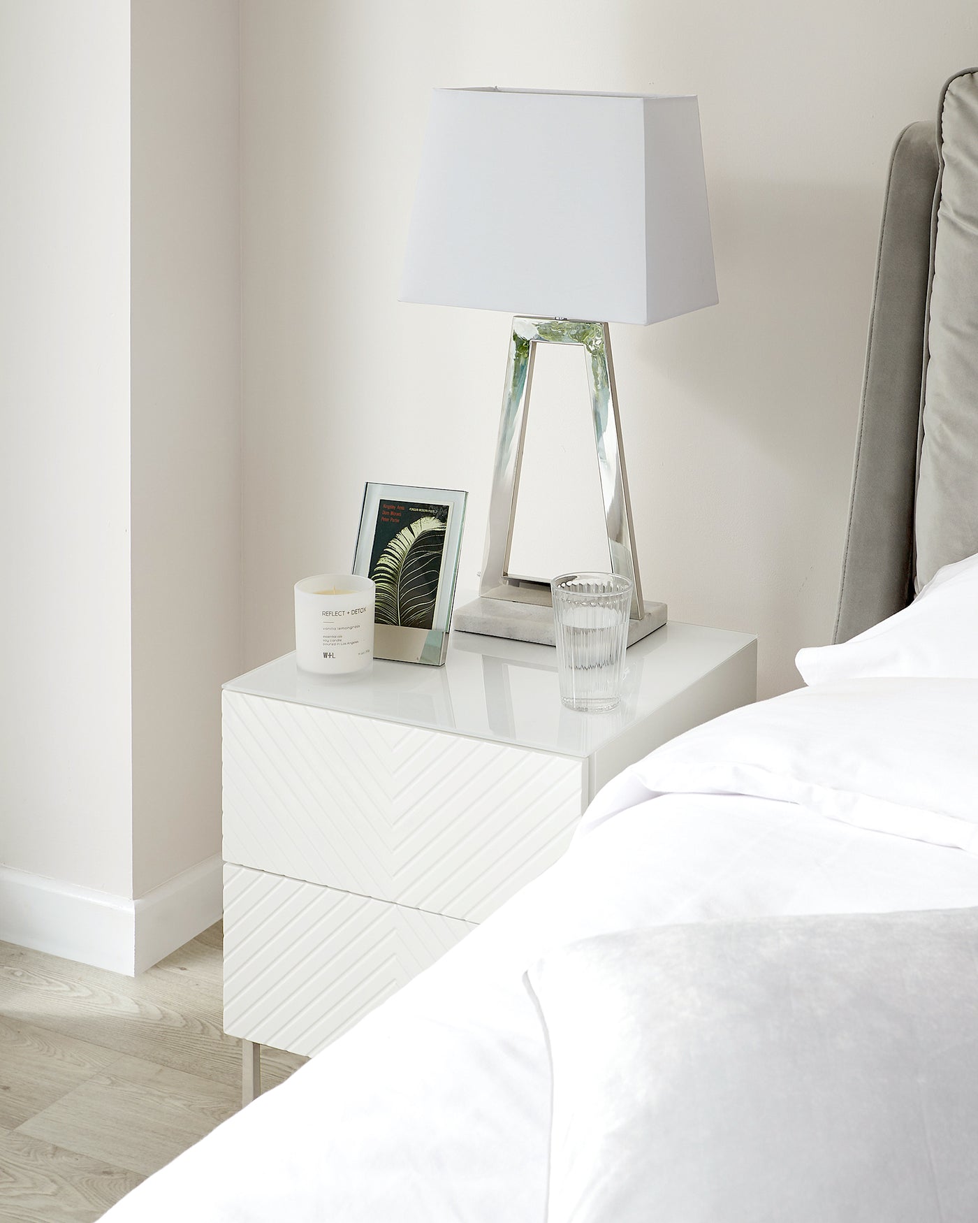 Modern white bedside table with a geometric patterned facade next to a bed with grey headboard.