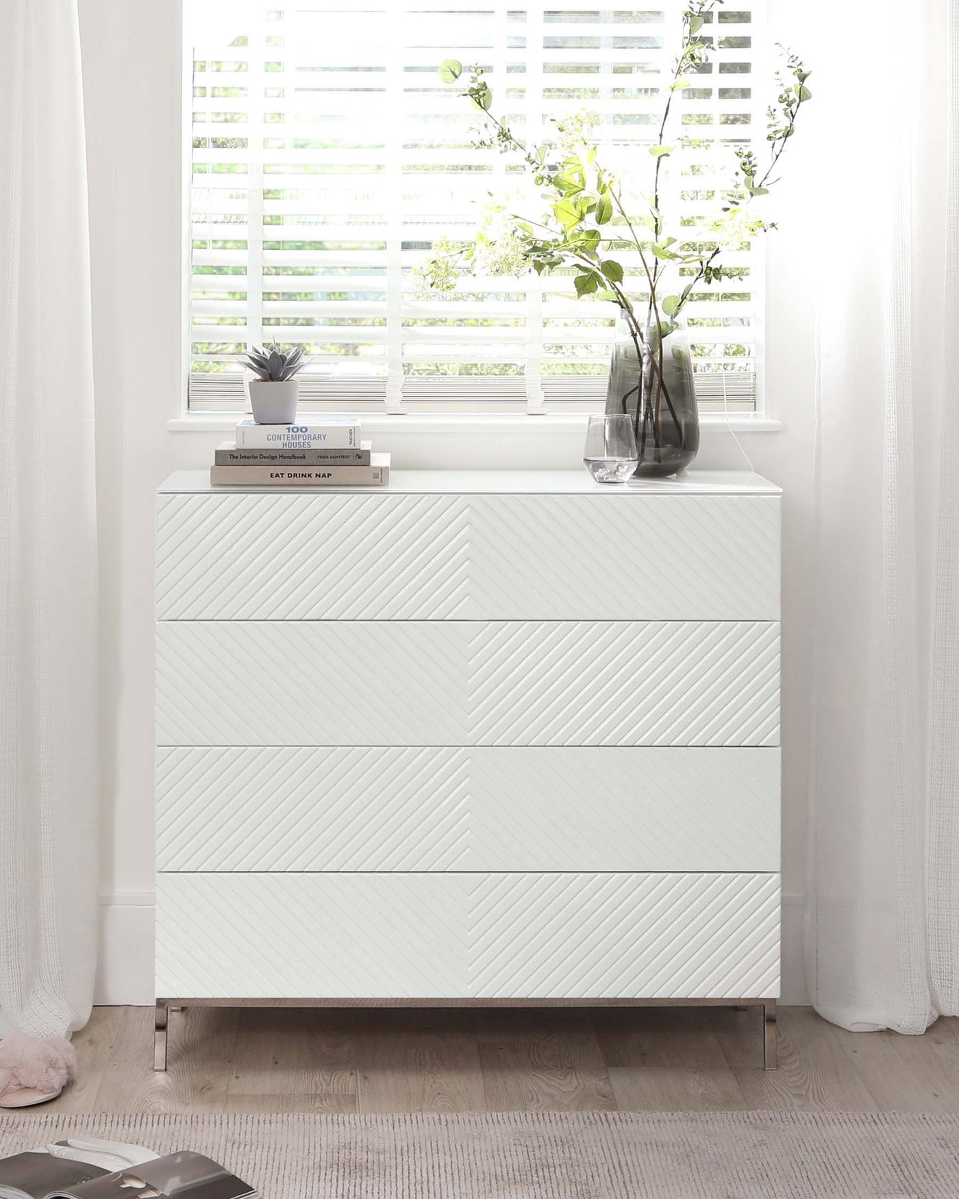 Modern white dresser with a textured chevron pattern on the drawer fronts and sleek metallic legs, accessorized with books, decorative plants, and a vase.