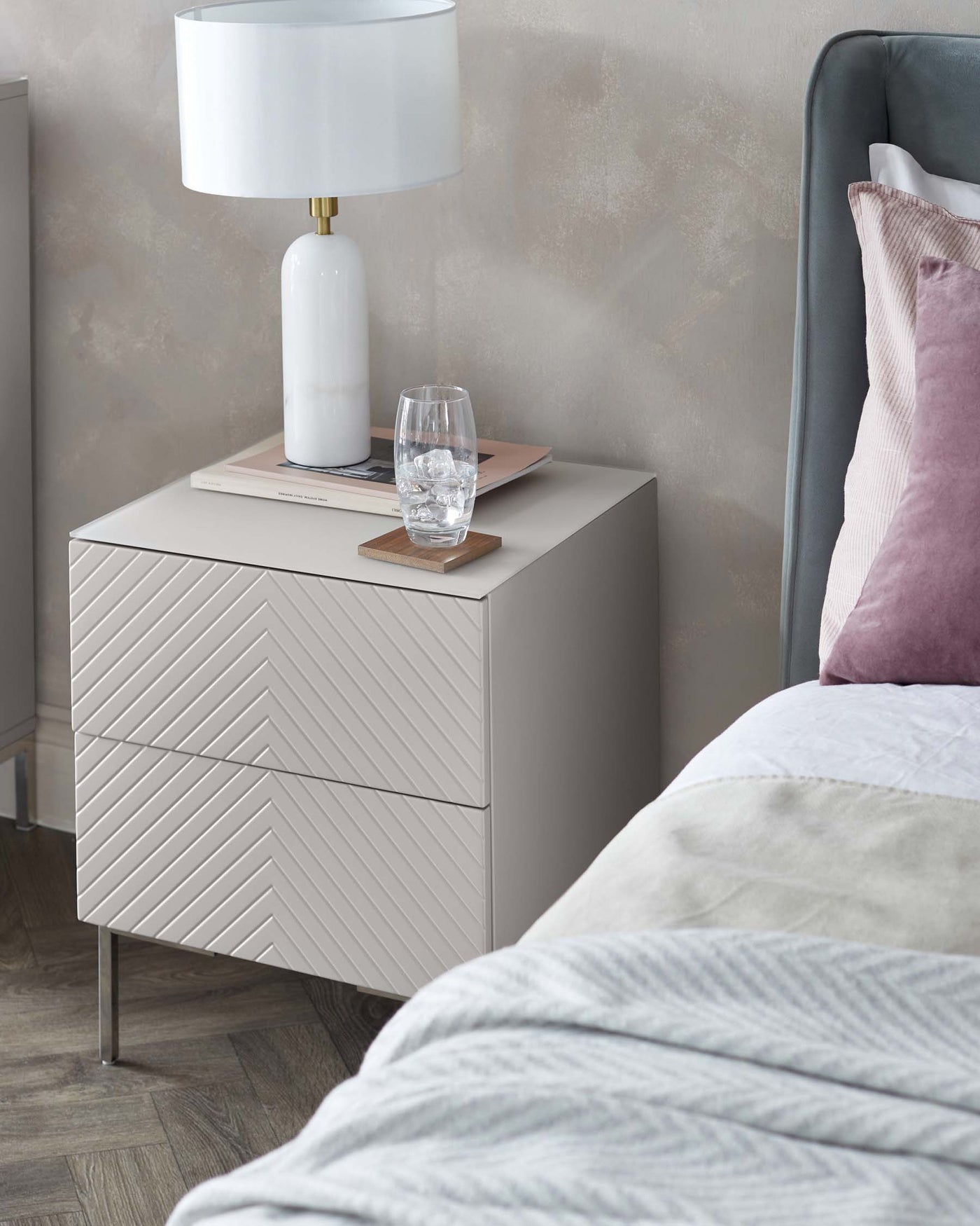 Modern light grey bedside table with chevron-patterned drawers and metal legs, accompanied by a white cylindrical lamp with a gold accent on top.