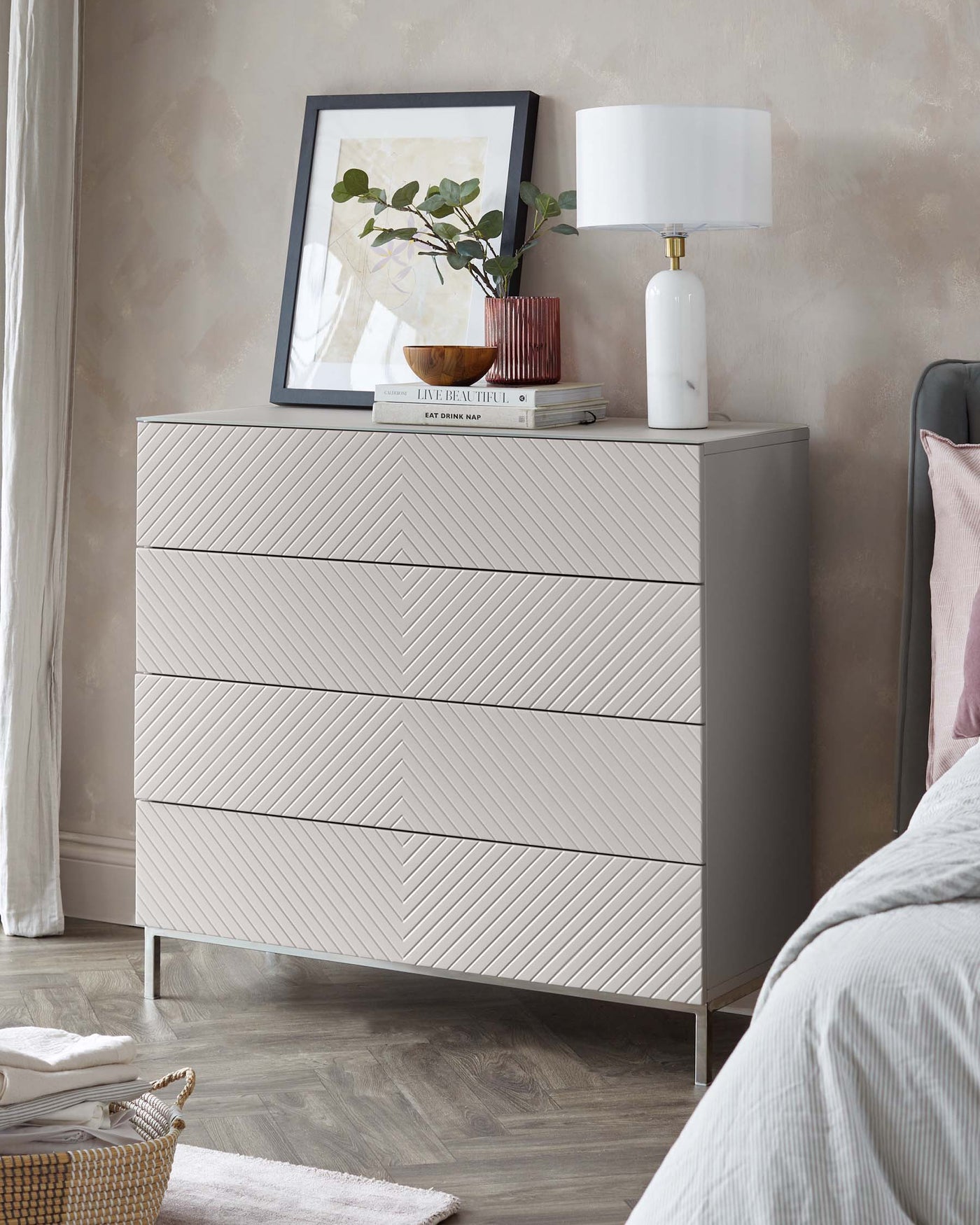 Modern light grey dresser with chevron patterned drawers and metal legs displayed in a bedroom setting with a lamp, framed artwork, and decorative items on top.