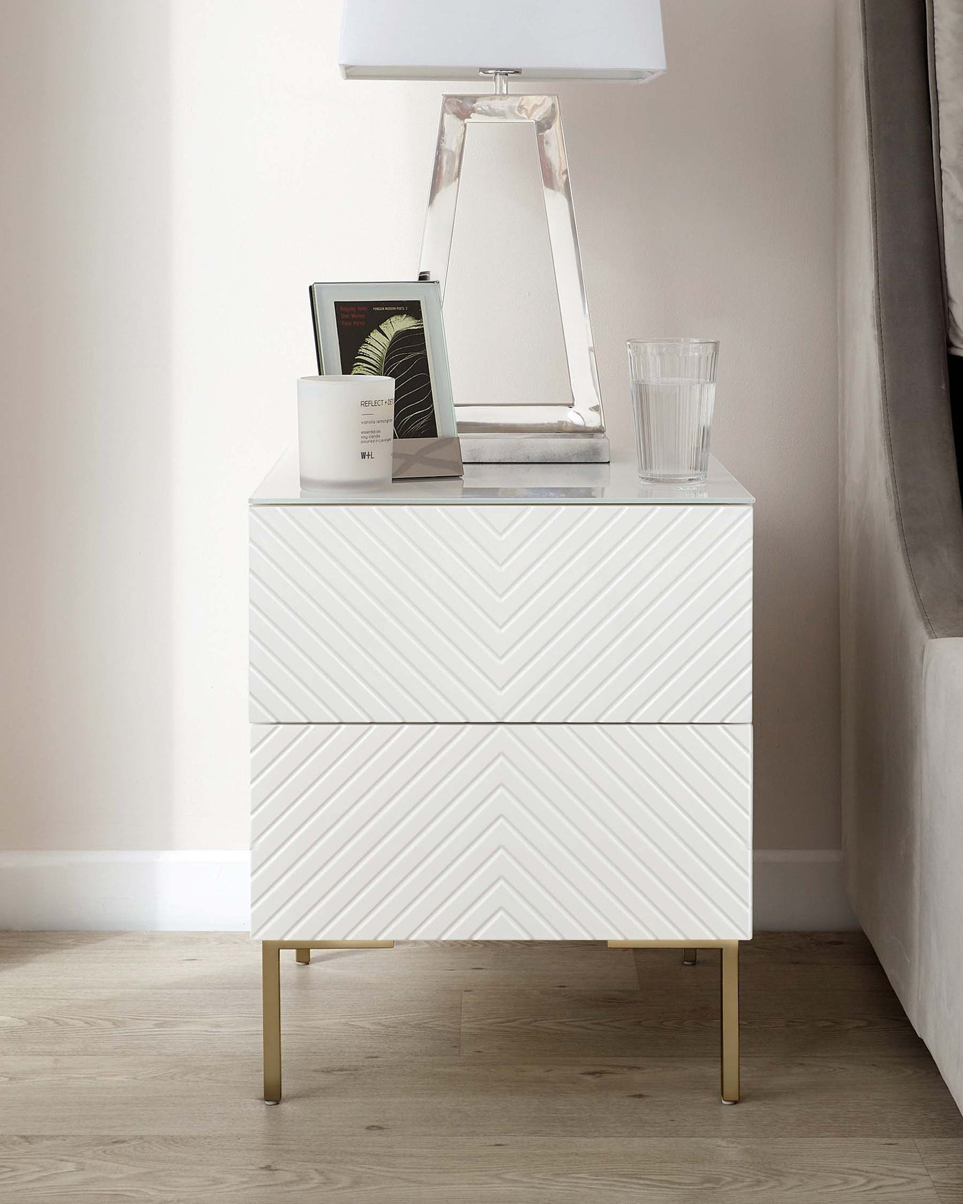 A contemporary white two-drawer nightstand with chevron patterned drawer fronts and brass-finished metal legs, adorned with a clear glass table lamp, a framed picture, and a clear water glass.
