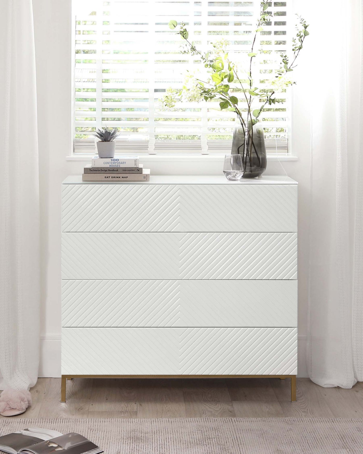 Modern white dresser with a textured front and brass-coloured legs, styled with a clear glass vase containing green branches, a small potted plant, and a stack of books.