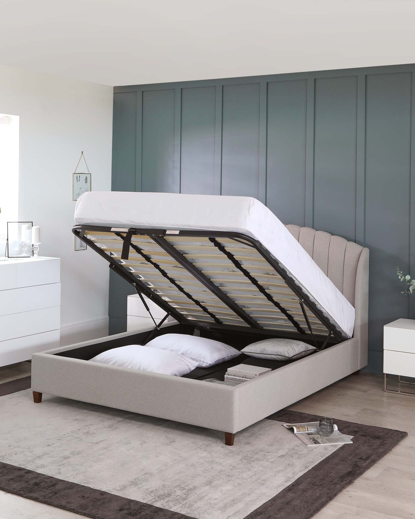 Upholstered lift-up storage bed with a tufted headboard and wood feet, featuring wooden slats partially visible under a raised mattress platform. A white nightstand with drawers and a clear glass lamp to the side; all staged on a grey area rug with a decorative clock and unlit candle as accents.