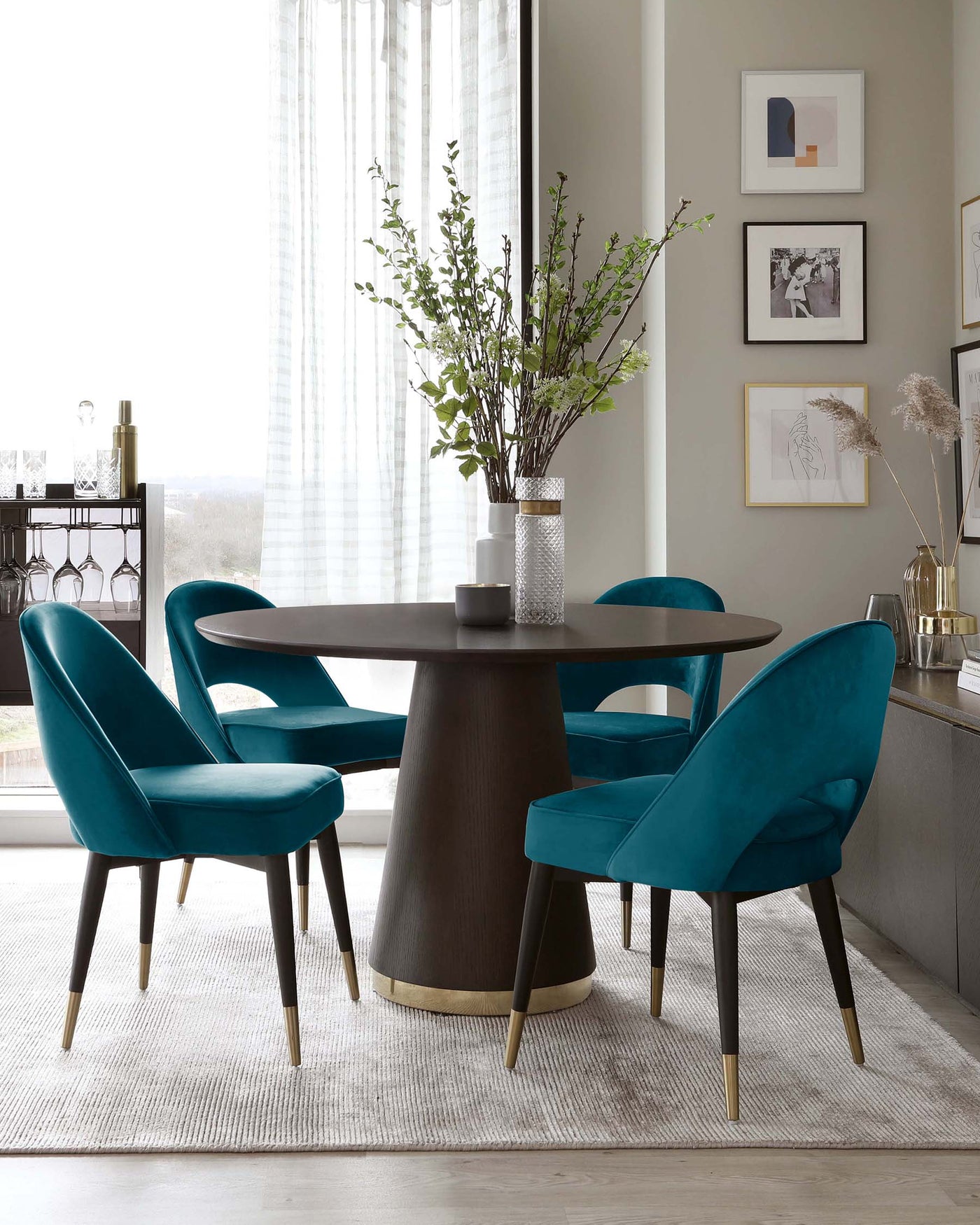 A modern dining set featuring a round, dark wooden table with a tapered base accented with a gold-coloured band at the bottom. Four plush velvet upholstered chairs in a rich teal colour with dark wooden legs tipped with gold-coloured accents encircle the table. The set sits atop a textured light beige rug.