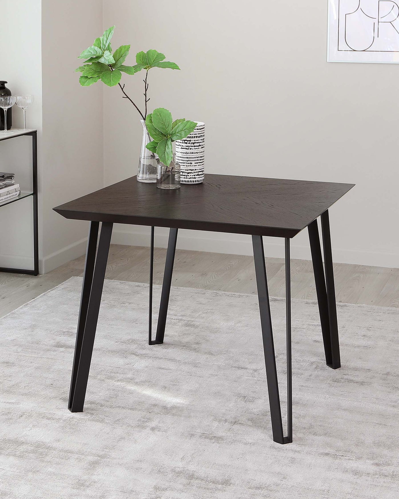 Modern minimalist dark wood dining table with angled black metal legs, displayed in a contemporary room with a light grey rug, accompanied by a clear glass vase with greenery and a decorative black and white patterned vase.