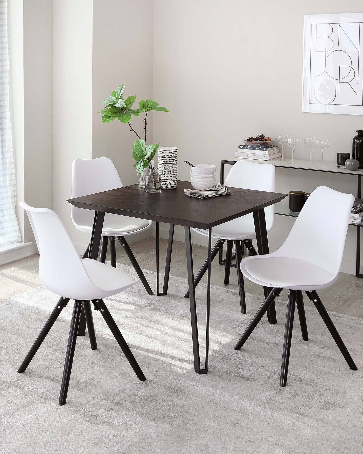 Contemporary dining set featuring a rectangular dark brown table with black tapered legs and four modern white chairs with black legs. The table is accessorized with a vase of greenery, books, and ceramic ware.