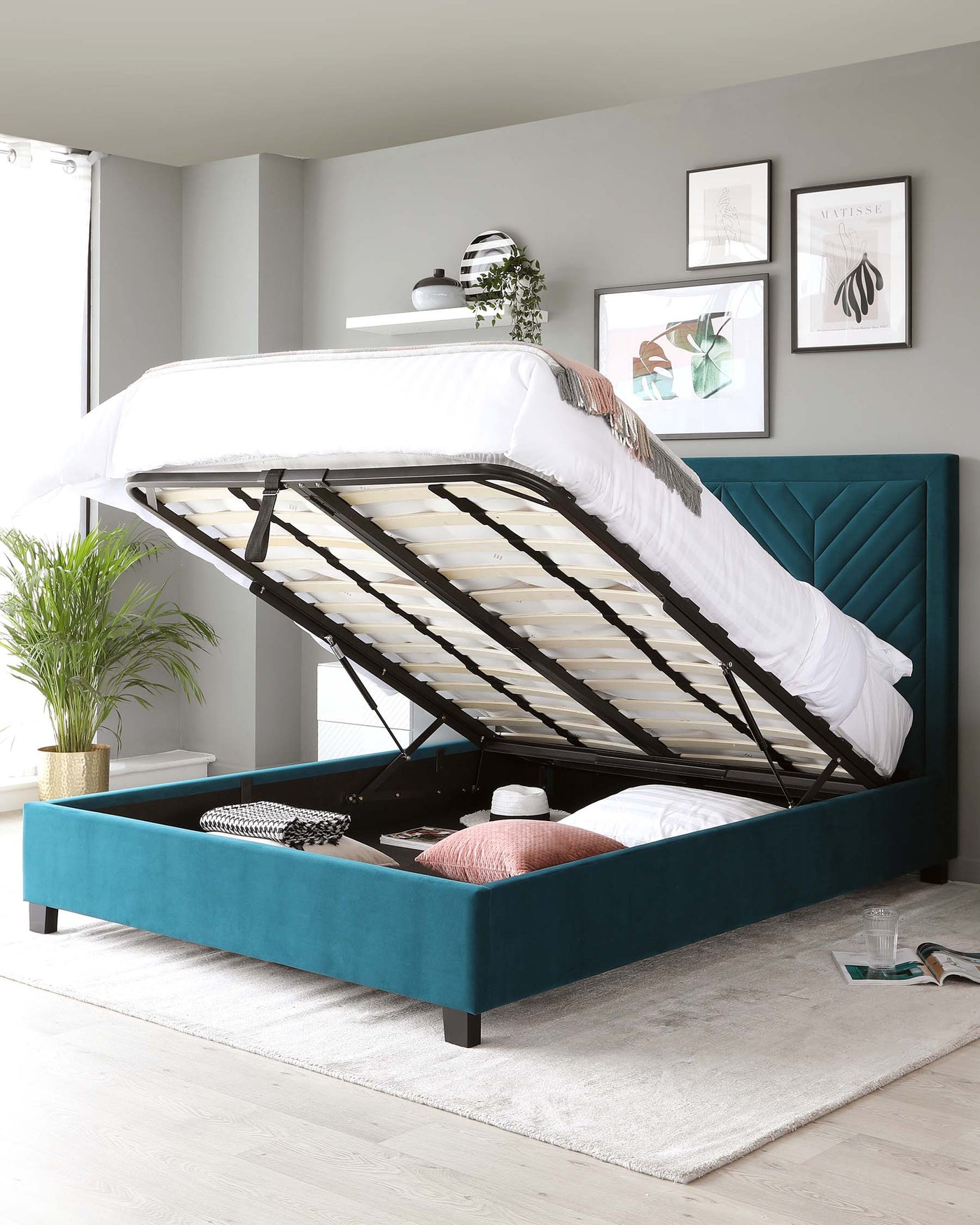 A contemporary teal upholstered storage bed with a lifted mattress revealing a spacious storage compartment underneath. The bed features a tufted headboard with a geometric design and is supported by dark wooden legs. A white mattress is partially visible, and beside the bed is a potted plant on a small stand. The room has a modern aesthetic with framed wall art and a minimalistic shelf above the bed.