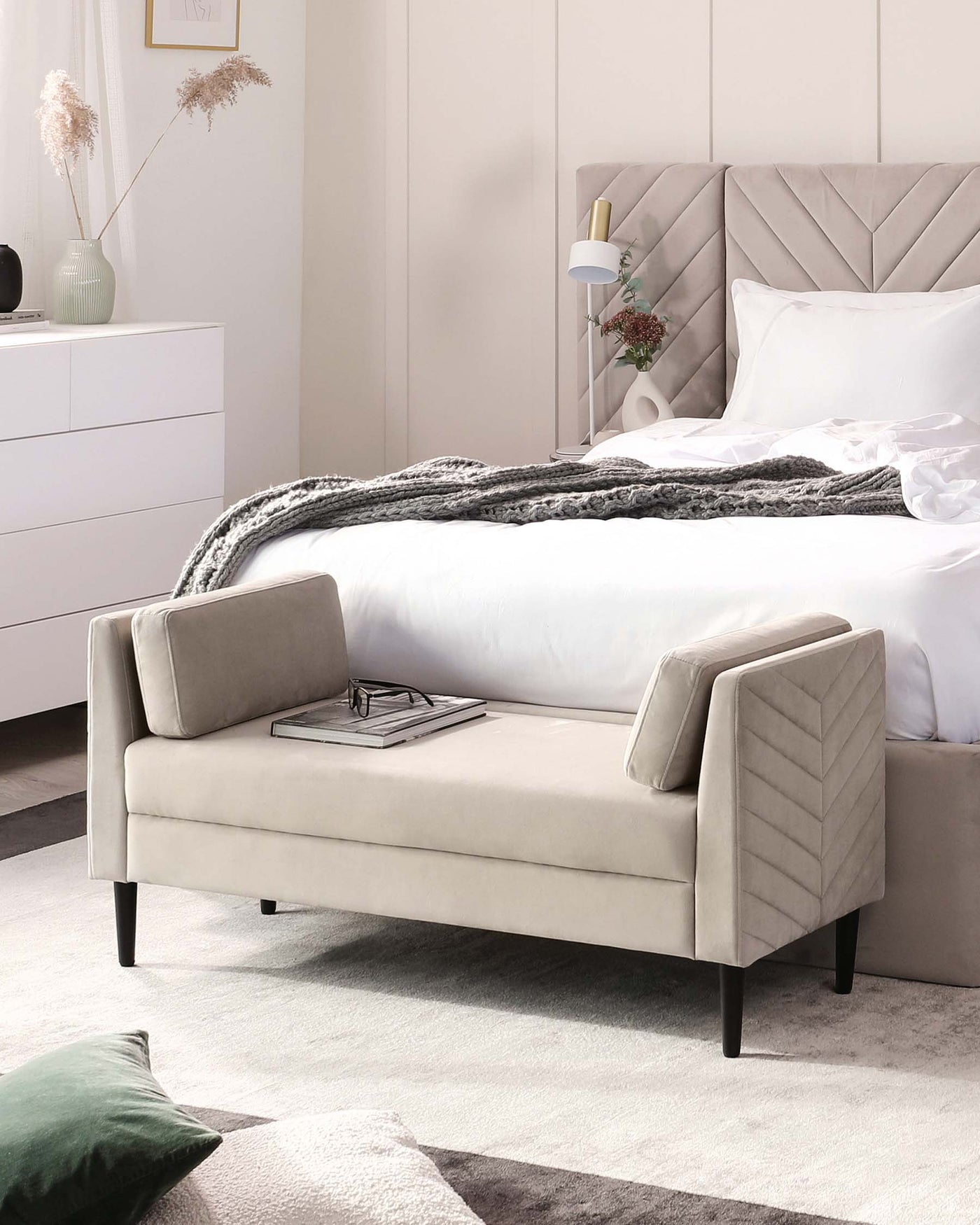 Elegant contemporary bedroom furniture set featuring a plush light grey upholstered bed with a diamond-patterned tufted headboard, flanked by a sleek white bedside dresser with minimalist handles. In the foreground, there is a matching light grey upholstered bench with clean lines and dark tapered legs, adorned with a simple decorative tray. The set is complemented by a soft-hued area rug, enhancing the room's modern and tranquil ambiance.