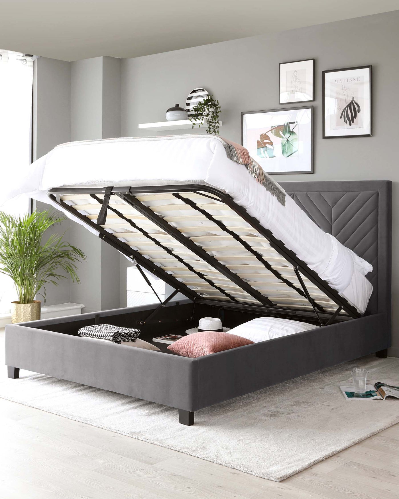 Contemporary grey upholstered storage bed with a gas-lift mechanism, revealing under-mattress storage compartment, tufted headboard, and black wooden feet, set in a modern bedroom with decorative elements.