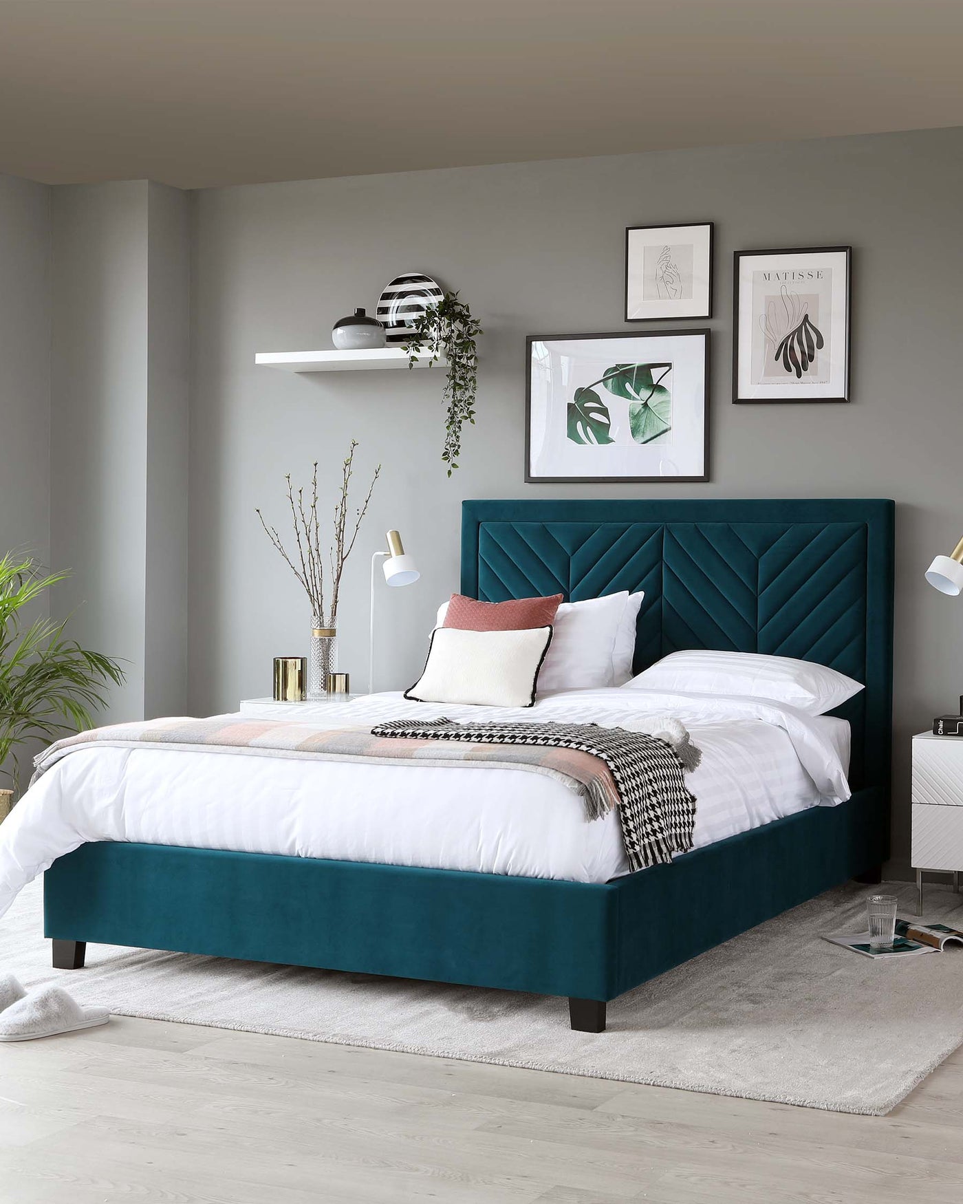 A luxurious teal upholstered bed with a tall, tufted headboard and a low-profile footboard, set on dark wooden legs. Coordinated bedding in white and shades of pink with a checkered throw complements the bed's rich colour. Two white minimalist bedside lamps provide symmetry, while a single floating shelf above the bed offers space for decorative items. The room is completed with a light grey area rug and two white bedside tables flanking the bed, enhancing the elegant and contemporary aesthetic.