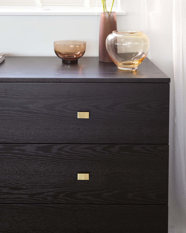 Modern black wooden dresser with three drawers, each featuring a sleek brass rectangular pull. The dresser's surface showcases a rich wood grain texture and is adorned with two decorative glass vases and a small bowl.
