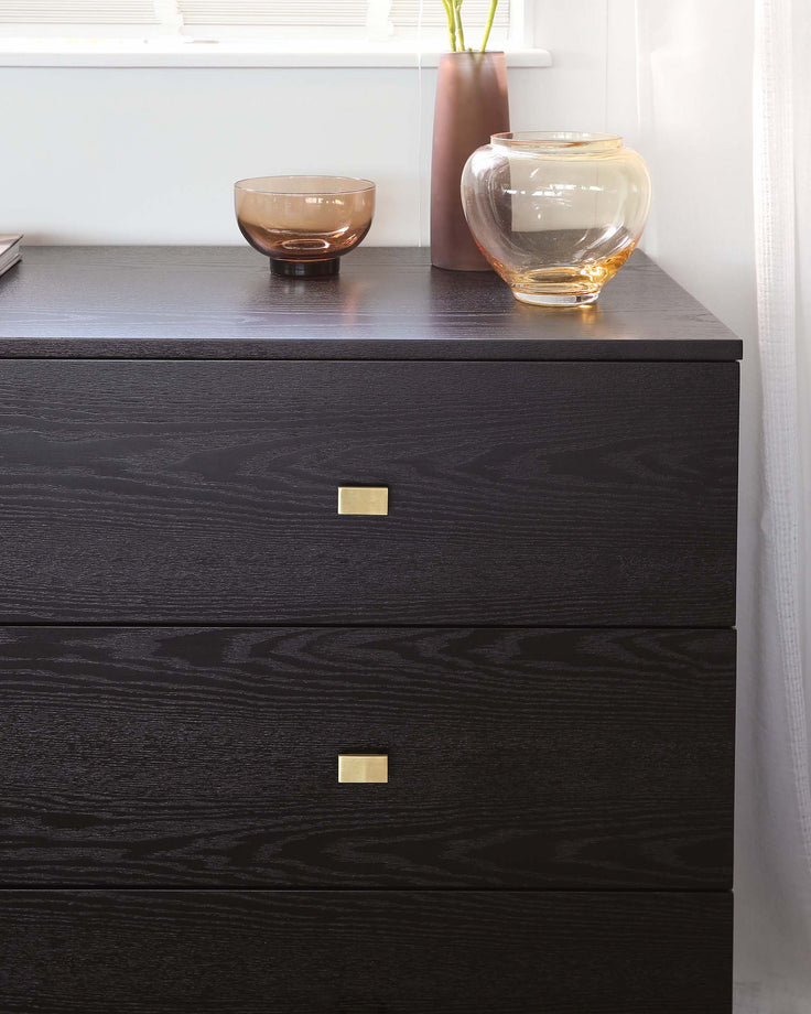 Modern black wooden dresser with three drawers, each featuring a sleek brass rectangular pull. The dresser's surface showcases a rich wood grain texture and is adorned with two decorative glass vases and a small bowl.