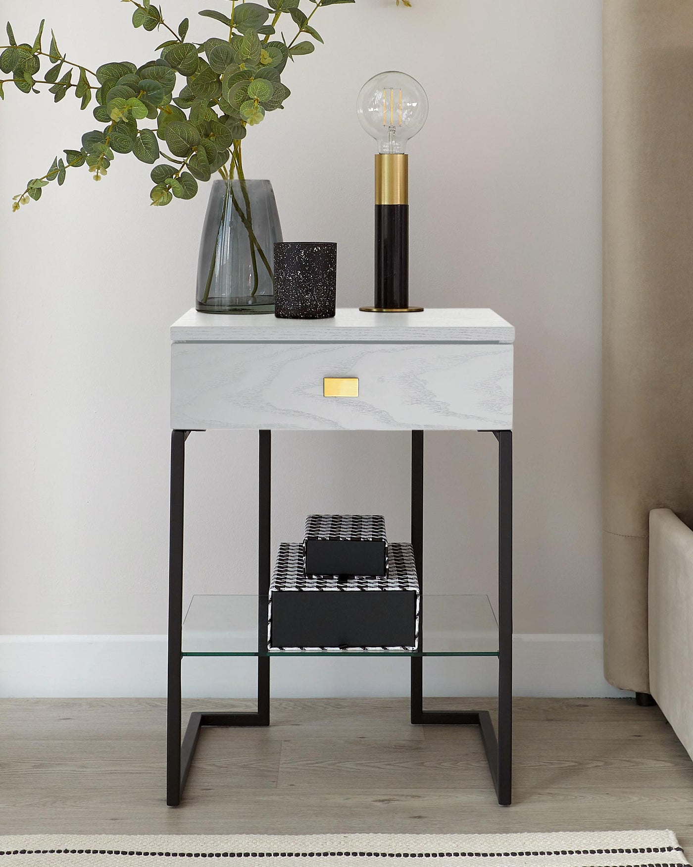 Modern marble-top side table with a sleek, minimalist design featuring a white and grey marbled surface, a single drawer with a gold accent handle, and a matte black metal frame. The table includes a lower shelf with a glass surface, perfect for displaying decorative items.