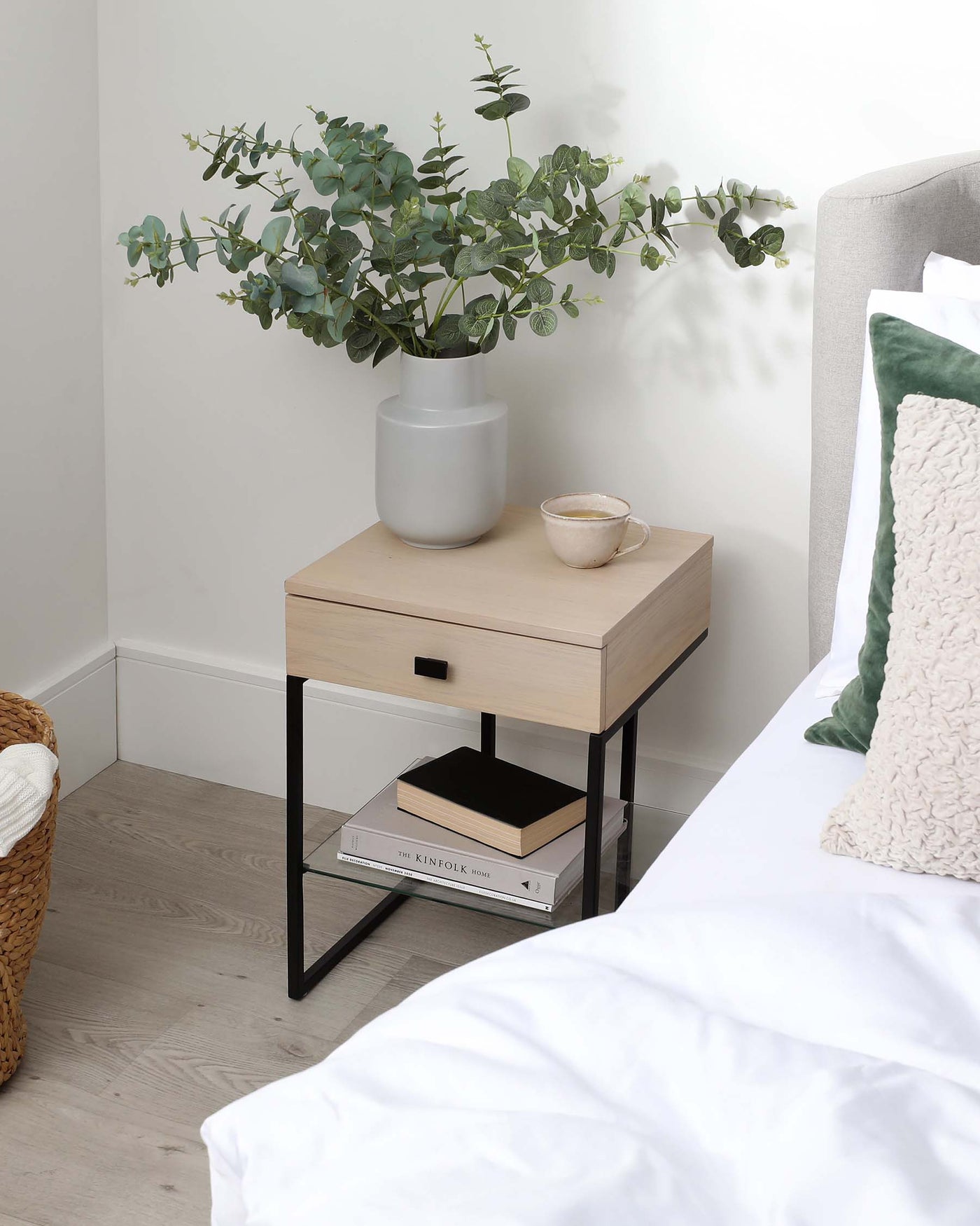 Modern minimalist bedside table with a light wood finish, featuring a single drawer with a cut-out pull handle and a lower shelf supported by a sleek black metal frame. The table is accessorized with a ceramic vase containing greenery and a small ceramic cup, accompanied by stacked books on the shelf.
