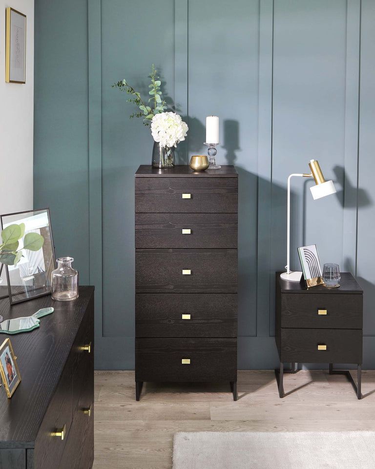 Modern dark wood finish furniture collection featuring a tall, narrow 5-drawer chest and a small 2-drawer nightstand, both with sleek brass handles and angled legs. A long sideboard with brass handles and leg accents partially visible on the left. The pieces are set against a teal wall and light hardwood floor, accessorized with vases, a lamp, and picture frames for a contemporary look.
