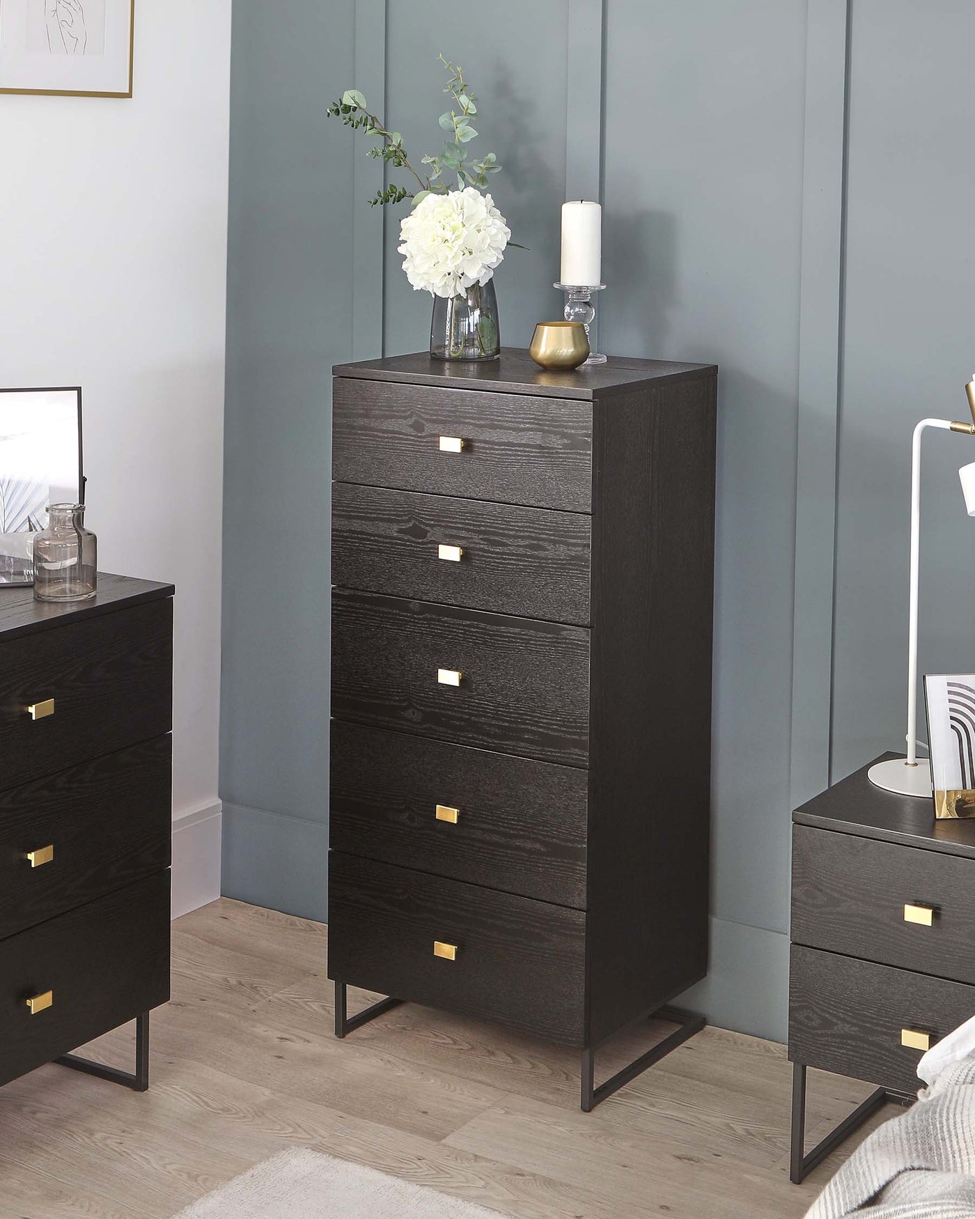 A collection of modern black wooden furniture featuring a tall 5-drawer chest and a shorter 3-drawer chest, both with gold-coloured metal handles and sleek, black metal legs. The furniture demonstrates a simple yet elegant design suitable for contemporary bedroom decor.