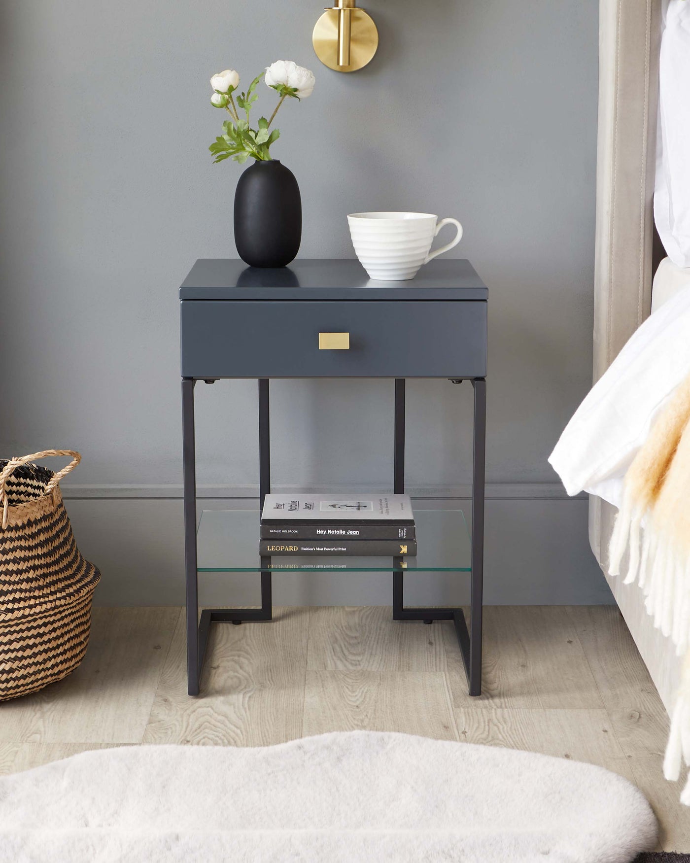 A modern dark grey nightstand with a single drawer, featuring a sleek brass handle and a lower shelf with a glass surface, supported by a minimalist metal frame. The nightstand is styled with a matte black vase holding white flowers and a white ceramic cup.