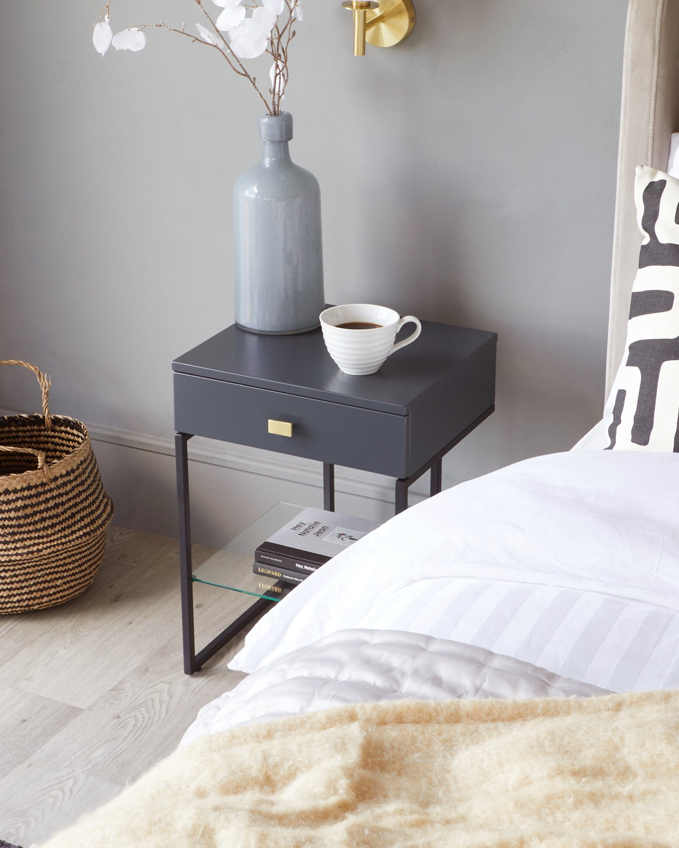 Modern dark grey bedside table with brass pull and lower glass shelf, beside a bed with neutral bedding.