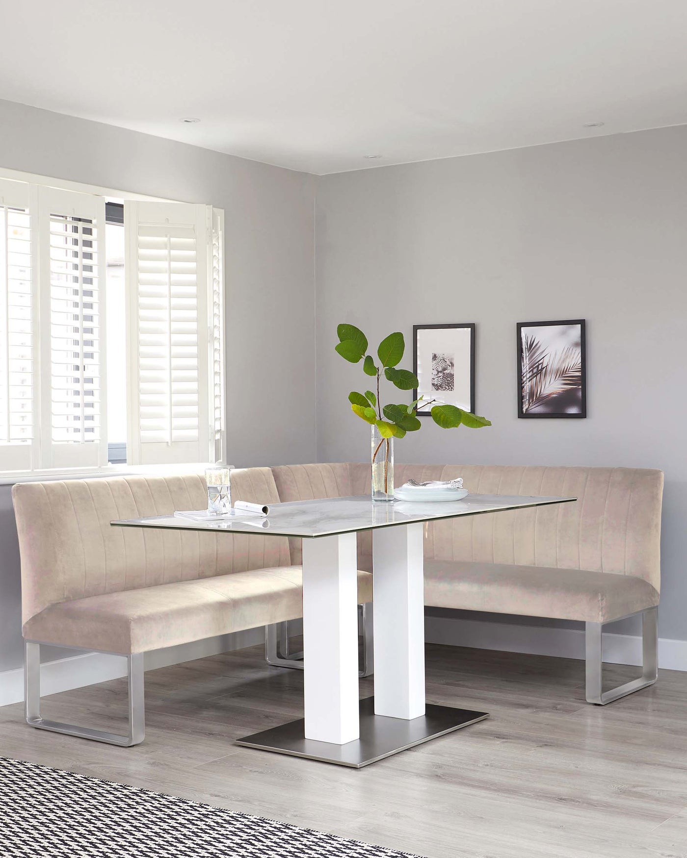 Modern dining room setup featuring a rectangular glass-top table with white and stainless steel legs, complemented by a matching L-shaped plush beige bench with back support, set against a neutral interior decor with wooden flooring.