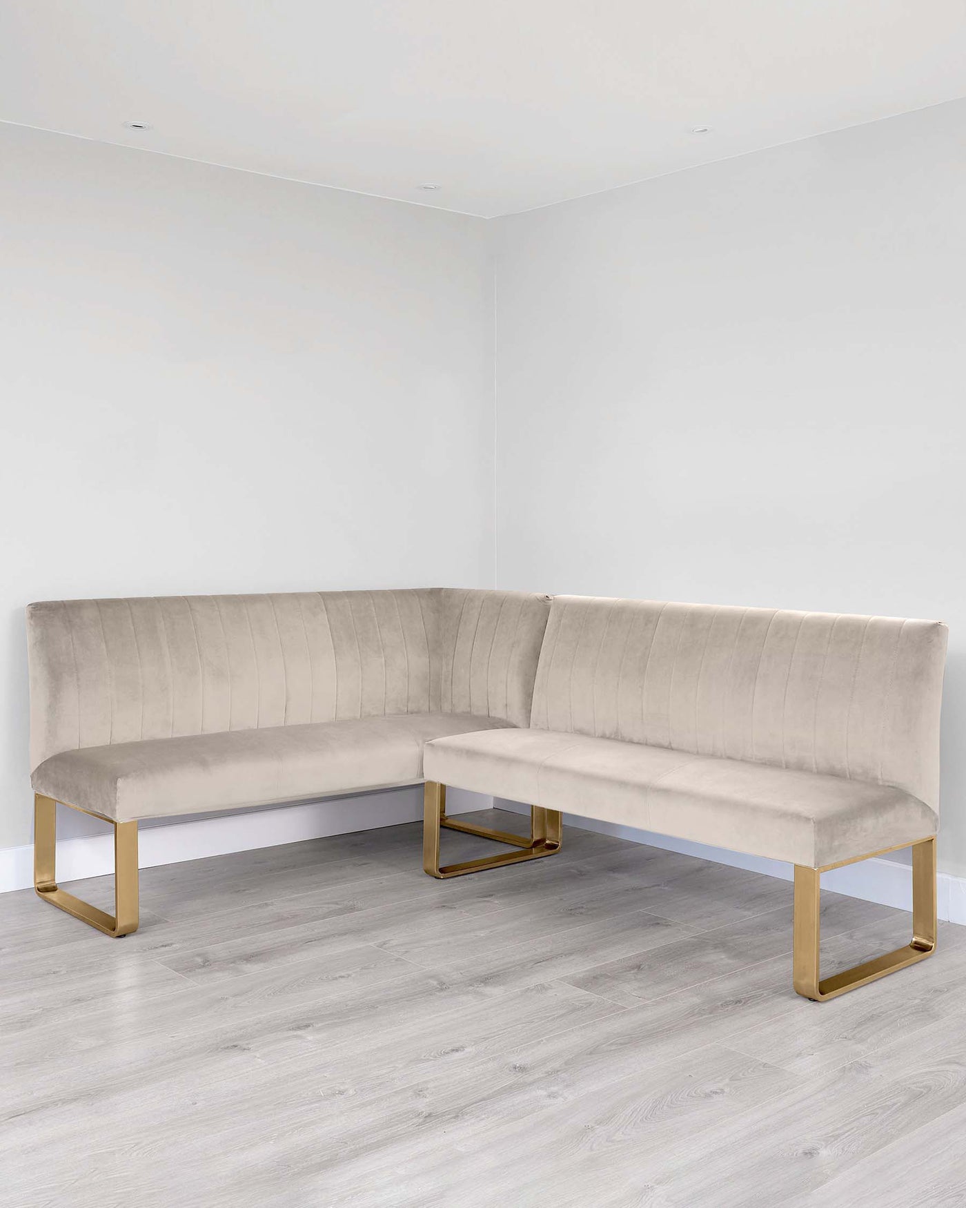 Modern L-shaped sectional sofa with velvet upholstery in a neutral colour, featuring a channel tufted backrest and sleek metallic gold legs. The sofa is presented in a minimalist setting with light wood flooring and white walls.
