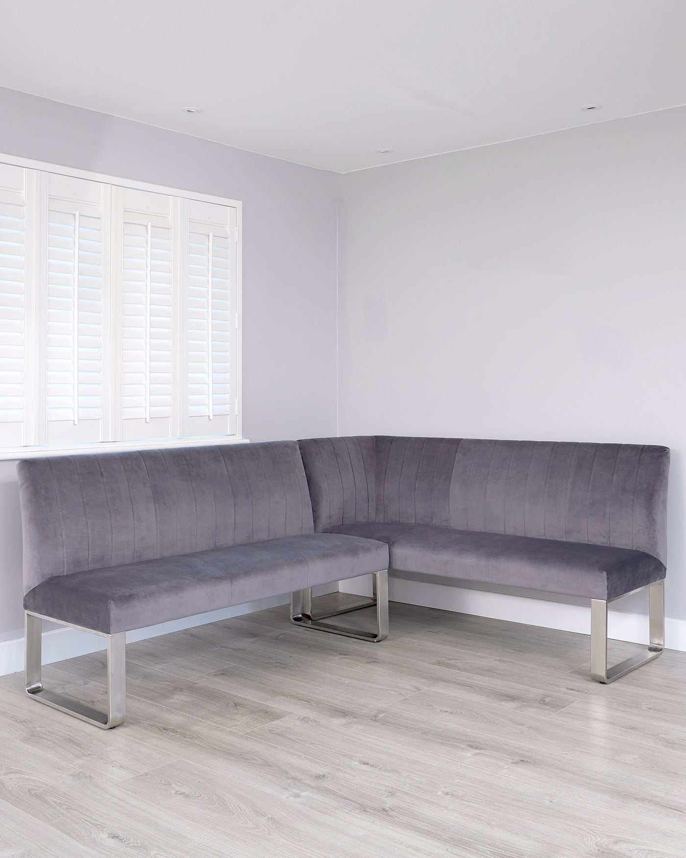 Modern L-shaped grey fabric corner sofa with clean lines and metal legs, set in a minimalist room with white plantation shutters and light wood flooring.