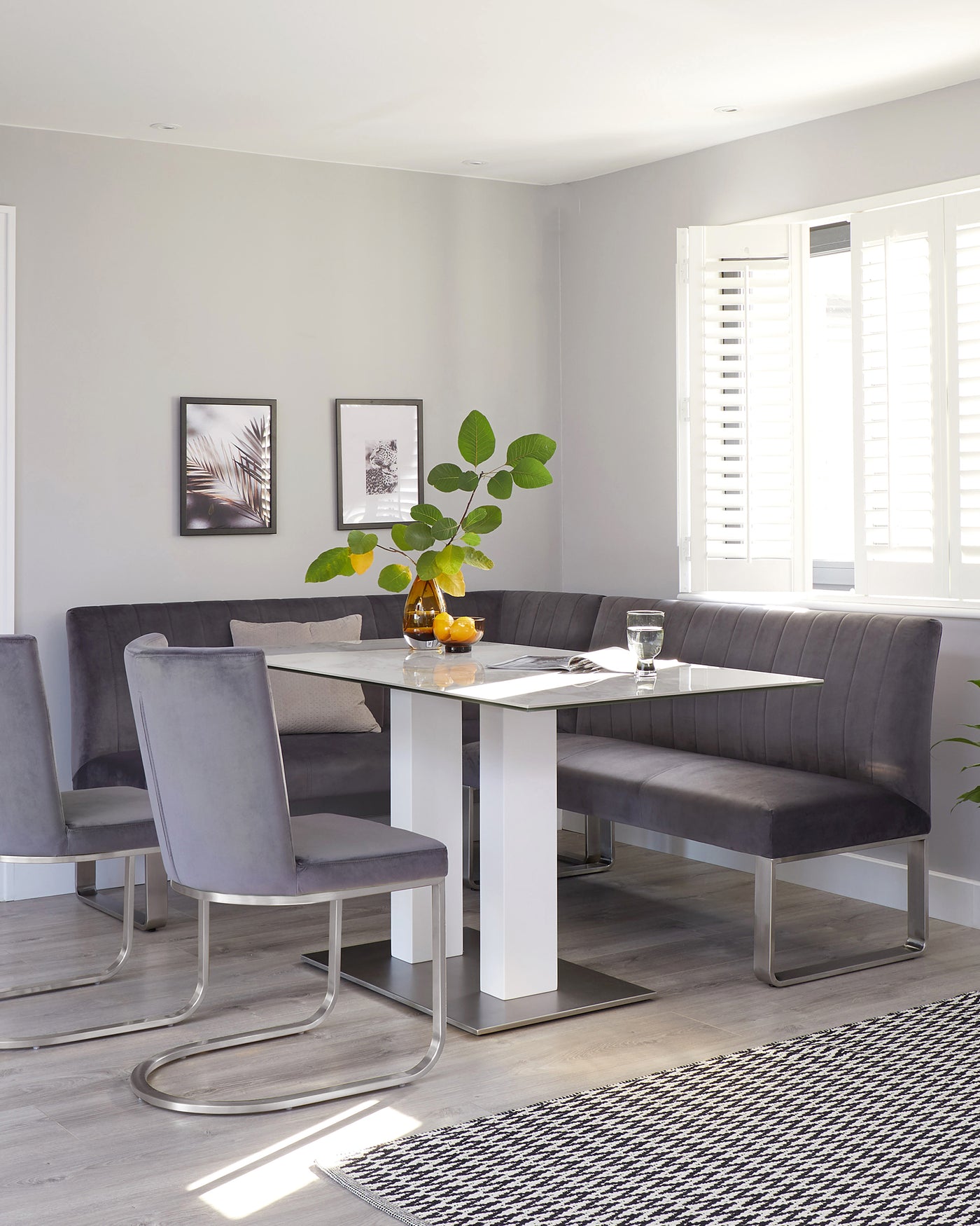 A contemporary dining set featuring a rectangular white table with a glossy finish and chrome accents at the base. Accompanying the table is a U-shaped, charcoal grey upholstered bench with a tufted backrest and three matching chairs with curved chrome legs and padded grey upholstery. The set is positioned on a sleek, wood laminate floor with a geometric black and white area rug beneath.