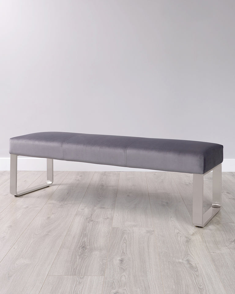 Modern long upholstered bench in charcoal grey with sleek chrome-plated metal legs, displayed on a light wooden floor with a plain white background.