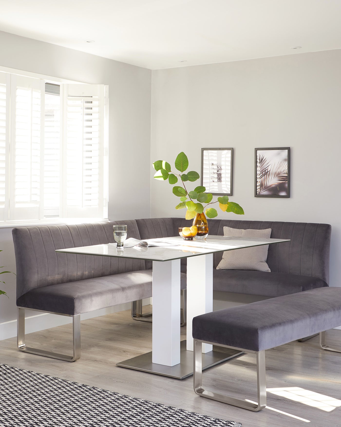 Modern dining room furniture consisting of a rectangular white table with a glossy finish and sleek metal legs, complemented by an L-shaped bench and a separate bench both upholstered in plush, grey fabric. The design features clean lines and a contemporary aesthetic.