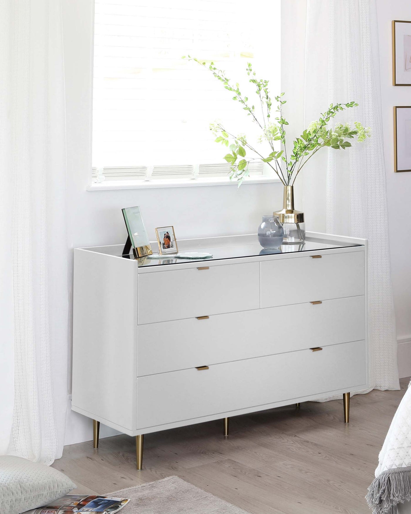 A modern white dresser with four drawers featuring slim horizontal handles and brass-coloured metal legs. The dresser is staged against a bright, airy room with a hardwood floor, and it is accessorized with decorative items including a brass vase with green foliage, a blue ceramic pot, and some picture frames.