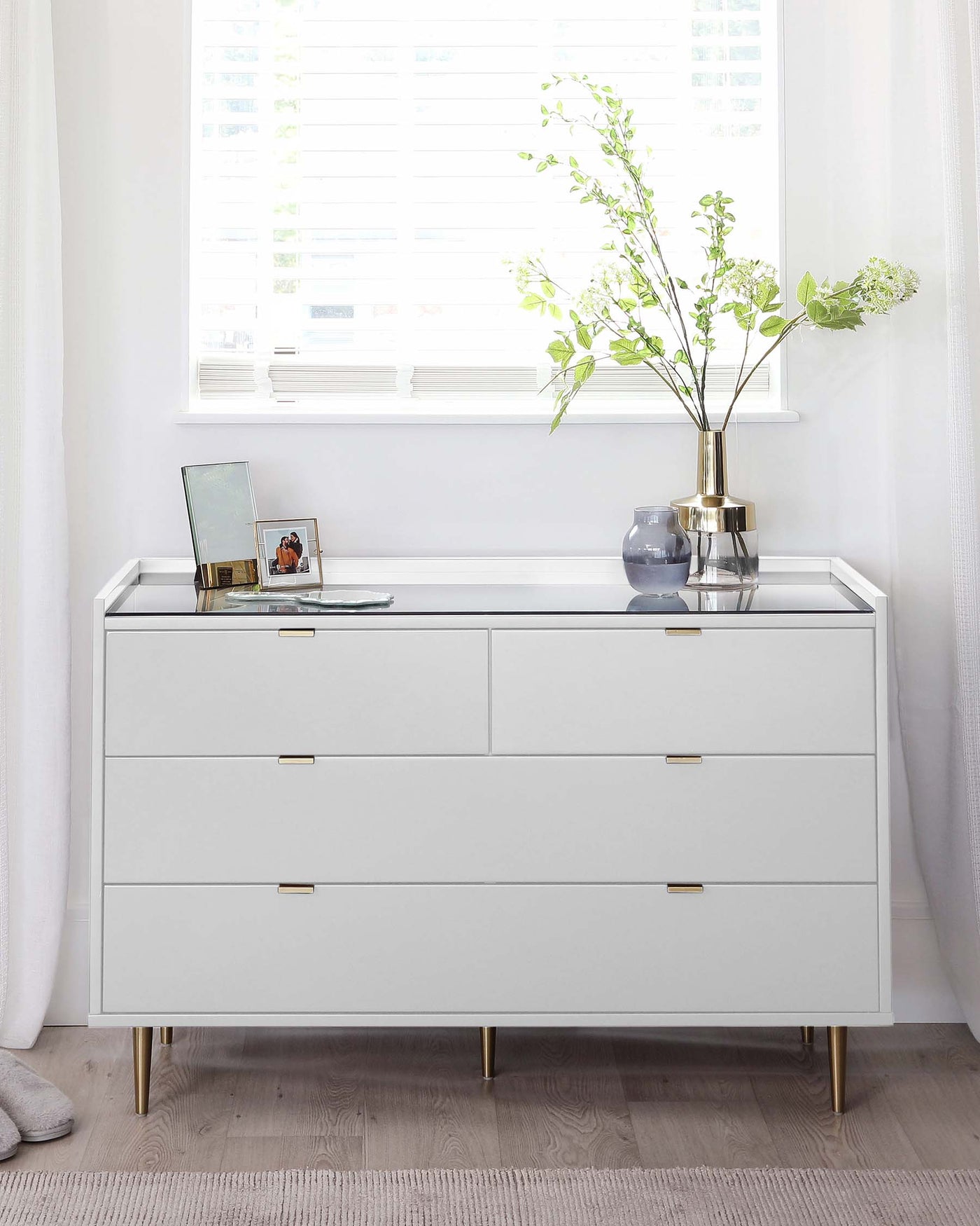 Modern white dresser with six drawers, featuring sleek brass handles and slender brass legs, against a light interior backdrop.