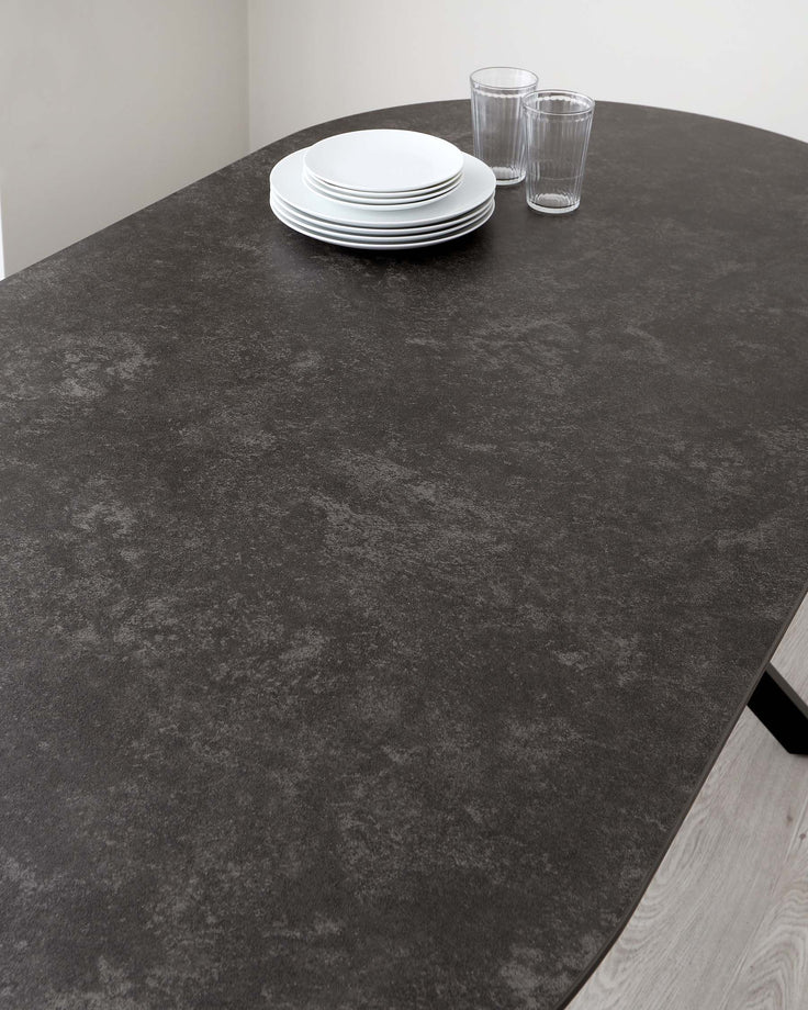 A contemporary round dining table with a dark, stone-texture finish on top and a light wooden base peeking out from one angle. The table is set with a stack of white dinner plates and two clear drinking glasses.