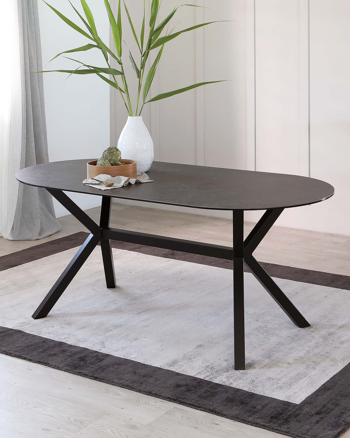 A modern oval-shaped coffee table with a dark grey matte top and angular black metal legs, displayed on a light grey area rug with a black border, in a bright room with white vertical panelling and light hardwood flooring.