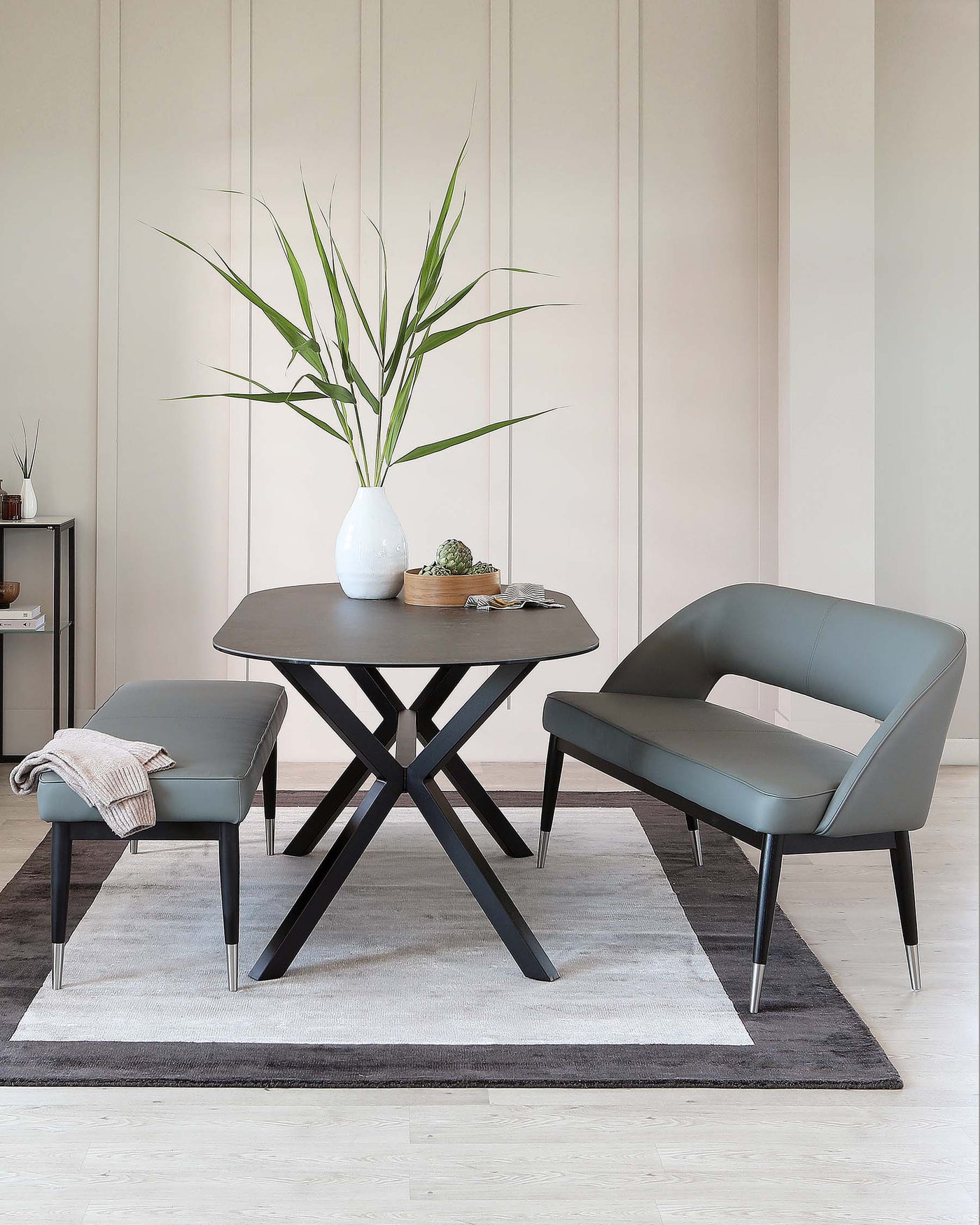 Modern minimalist furniture set in a neutral-toned room, featuring a sleek, round, black dining table with a unique X-shaped base, complemented by a pair of streamlined, dark grey armchairs with metallic accents on the legs. The set is arranged on a two-tone grey area rug, and the table is accessorized with a tall white vase containing green foliage, alongside small decorative items. In the background, a dark shelving unit holds assorted decor.