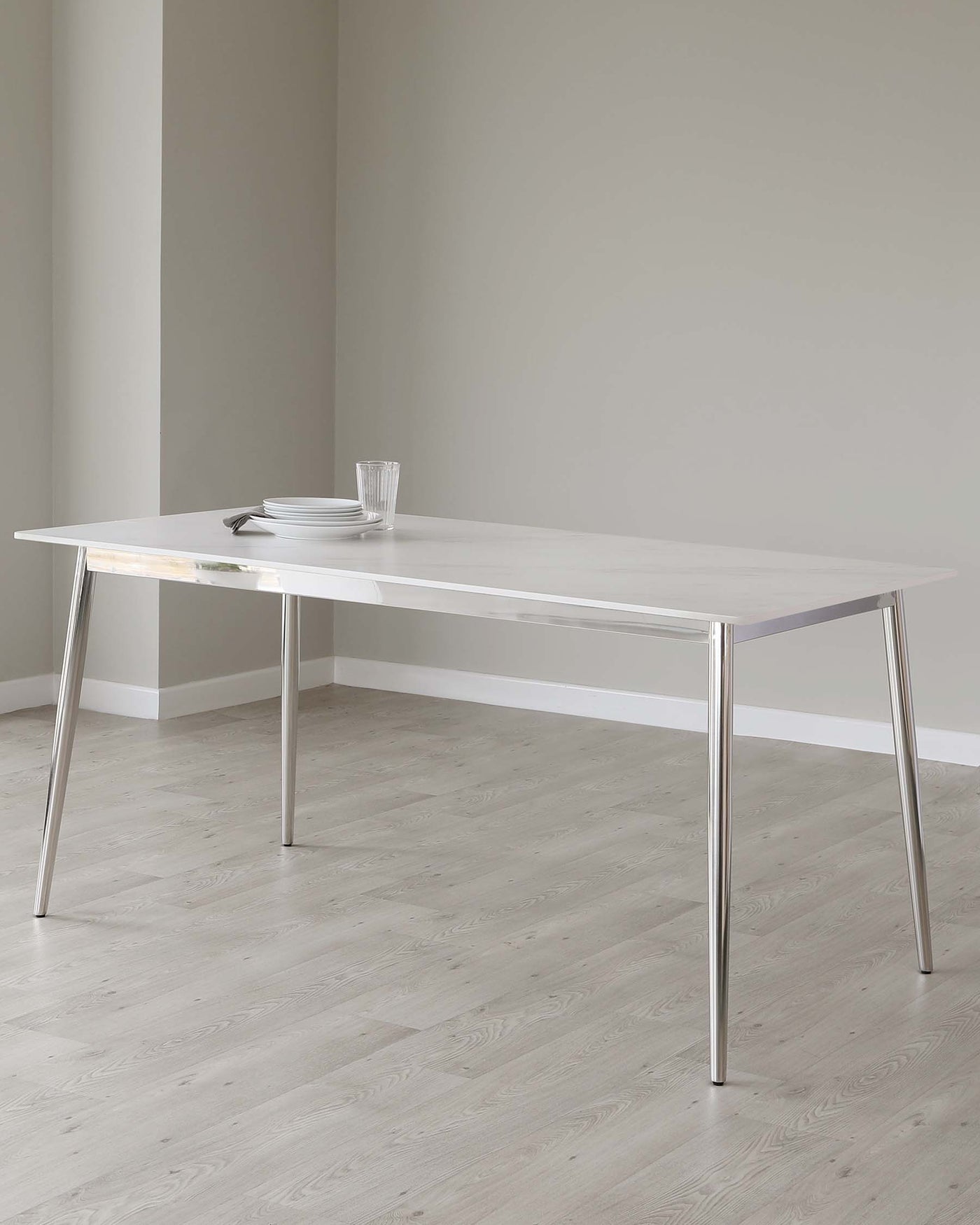 A modern minimalist white dining table with slender silver metal legs, set in a room with light grey walls and pale wooden flooring. The table is adorned with a simple set of white plates, a clear glass, and a pair of silver cutlery.