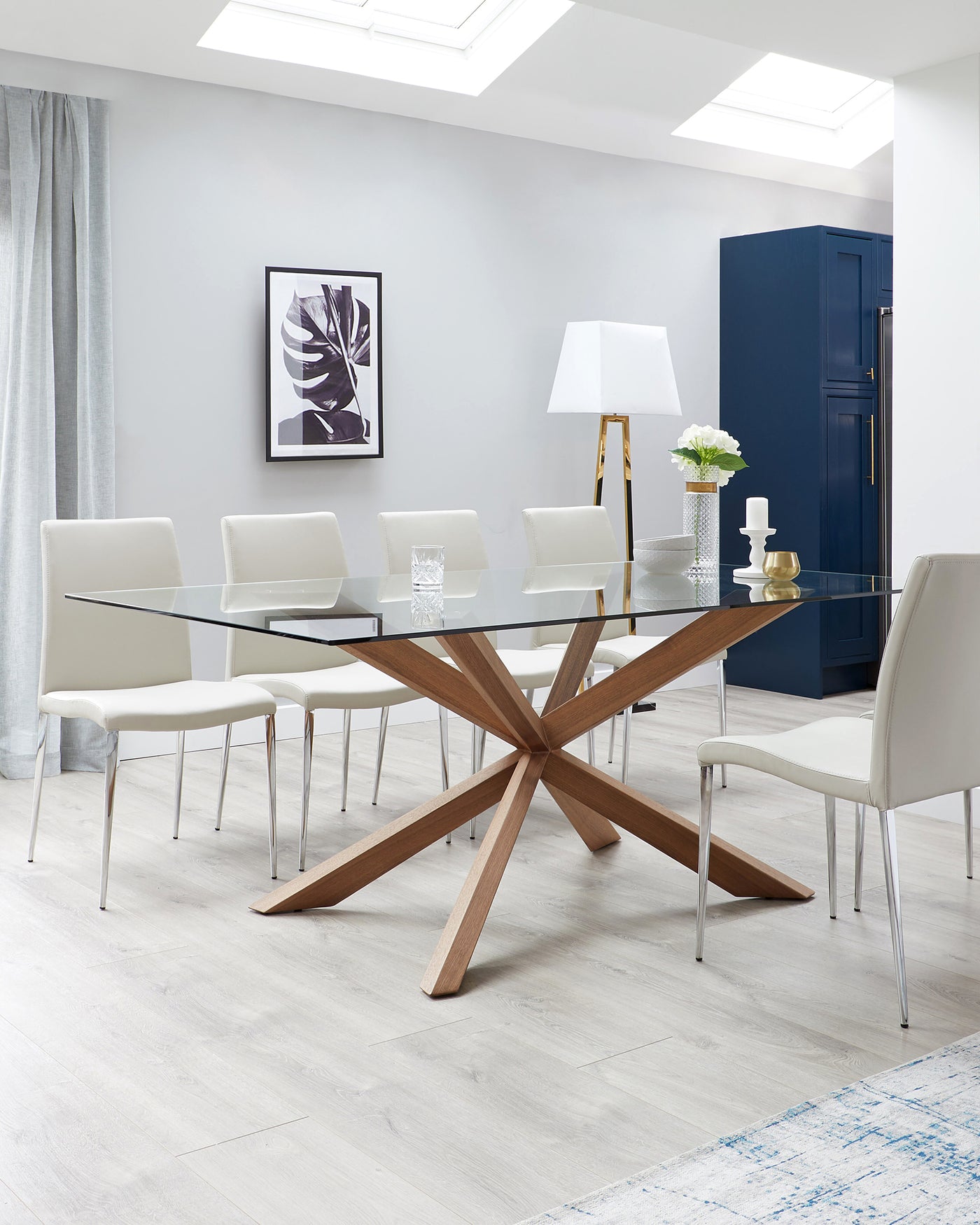 A modern dining room with a rectangular glass-topped table supported by a wooden starburst-style base. Surrounded by six sleek white leather chairs with slim metal legs. A stylish floor lamp with a brass stand and a white shade adds elegance to the corner of the room.