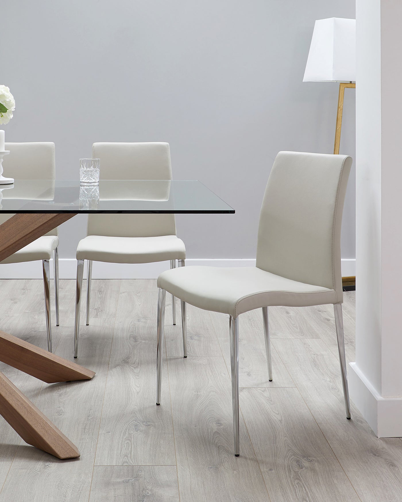 Modern dining set with a rectangular glass-top table on a wooden frame, surrounded by four sleek, white upholstered chairs with metal legs. A minimalist white floor lamp stands in the background.